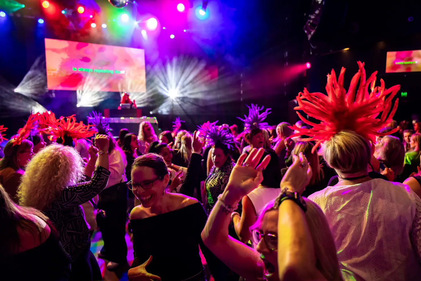 A crowd of ladies dance in front of the stage and bright stage lights, some wear feathered headbands