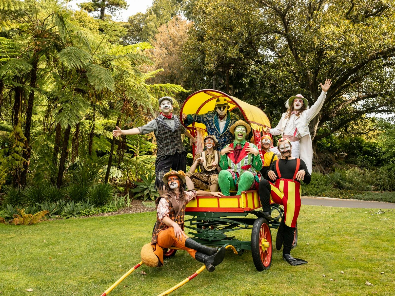 Cast in animal costumes performing on a yellow and green wagon, set against a garden backdrop.