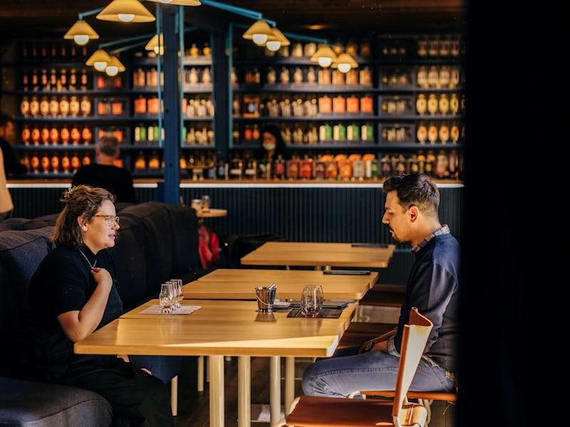 2 people sitting at a table having a whisky tasting with a bar and bottles in the background