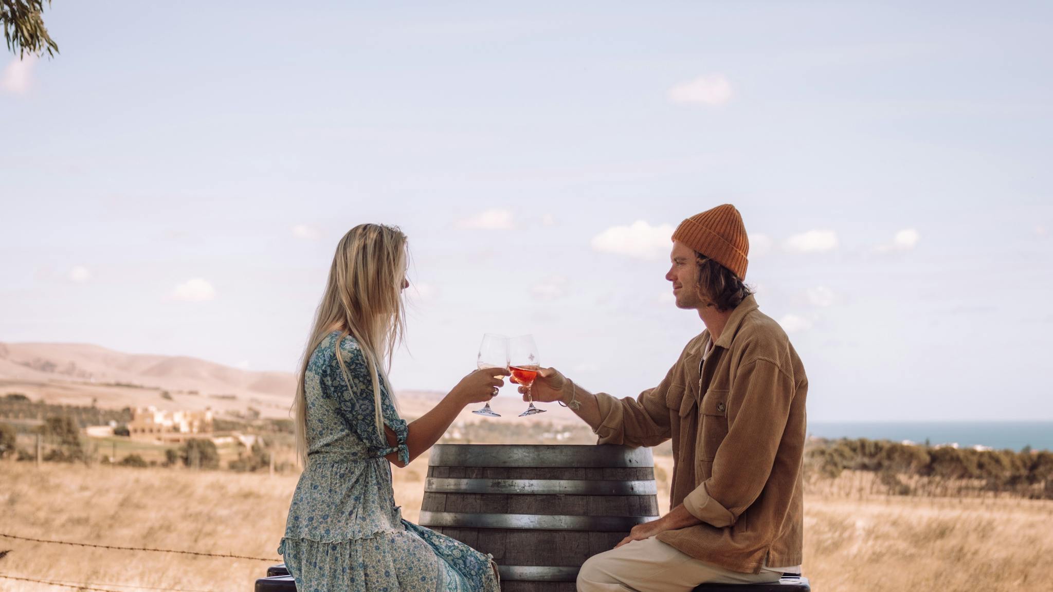 Two people holding wine, sitting at a wine barrel overlooking the hills and ocean