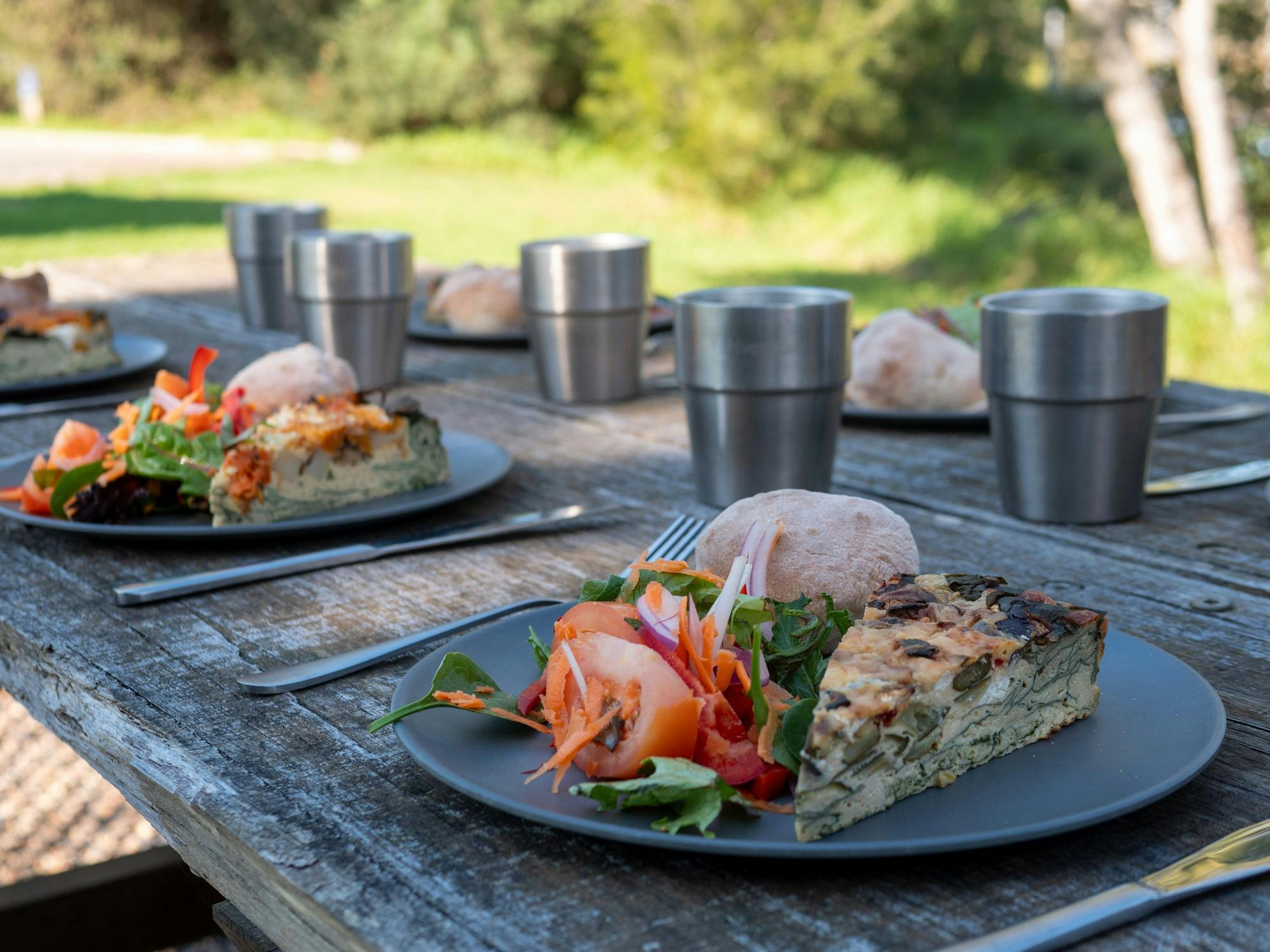 A picnic lunch with salad, frittata, bread roll, and a drink, set outdoors in nature.