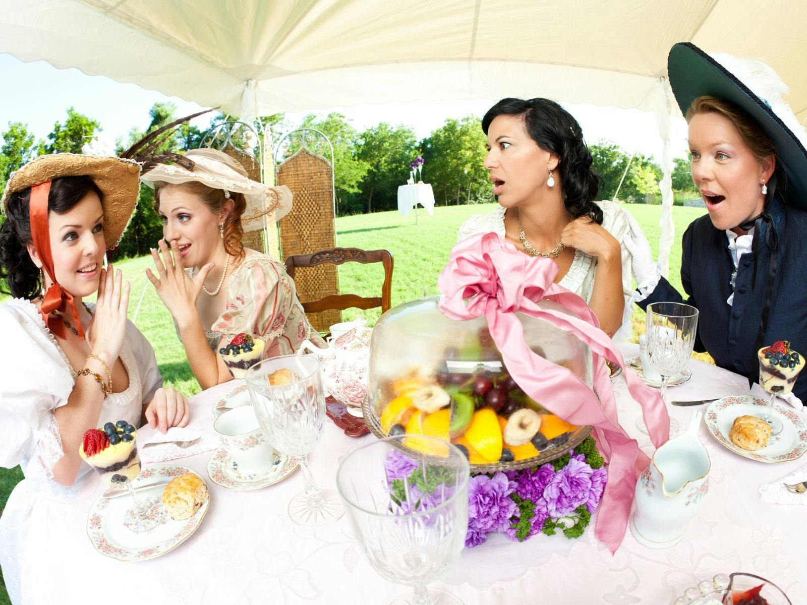 For women in historic dress sit gosspining at afternoon tea table