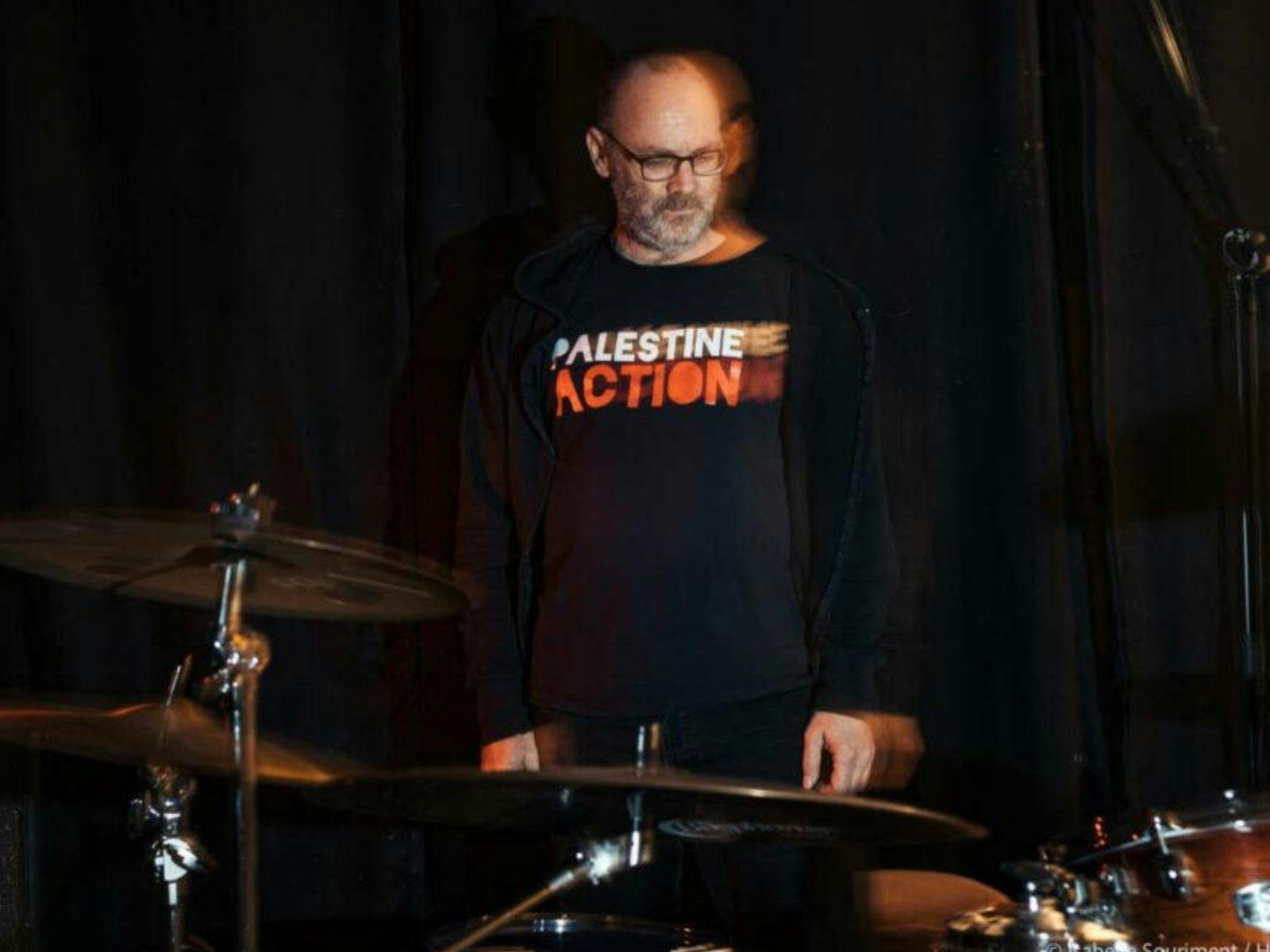 David Rovics standing behind drums with a Palestine Action t-shirt on. Black background.