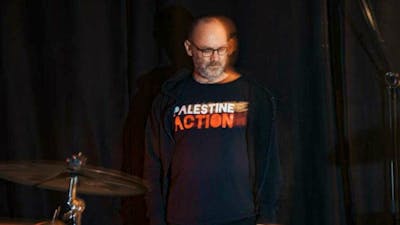 David Rovics standing behind drums with a Palestine Action t-shirt on. Black background.