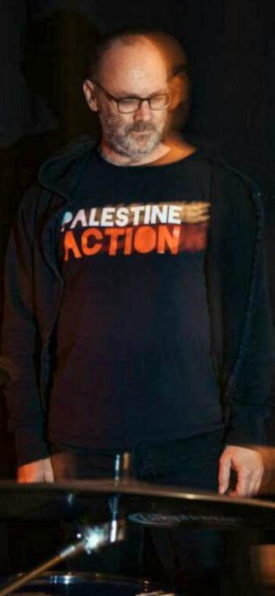 David Rovics standing behind drums with a Palestine Action t-shirt on. Black background.
