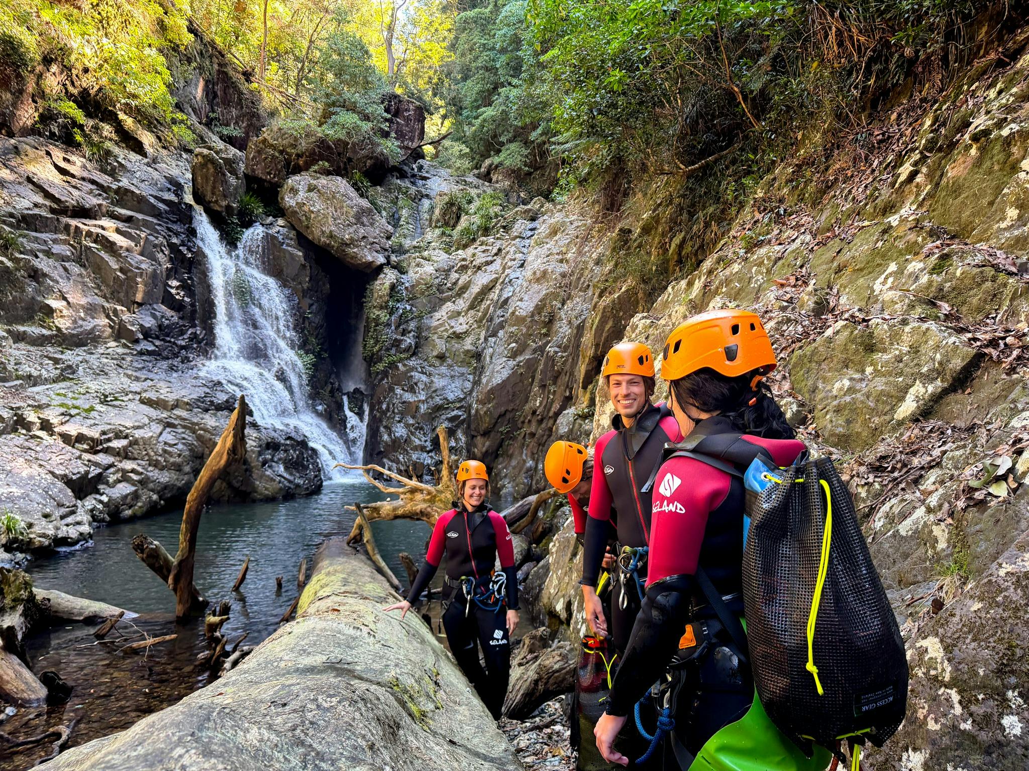 Group of canyoners in wetsuits and orange helmets beside a waterfall and deep pool in a canyon