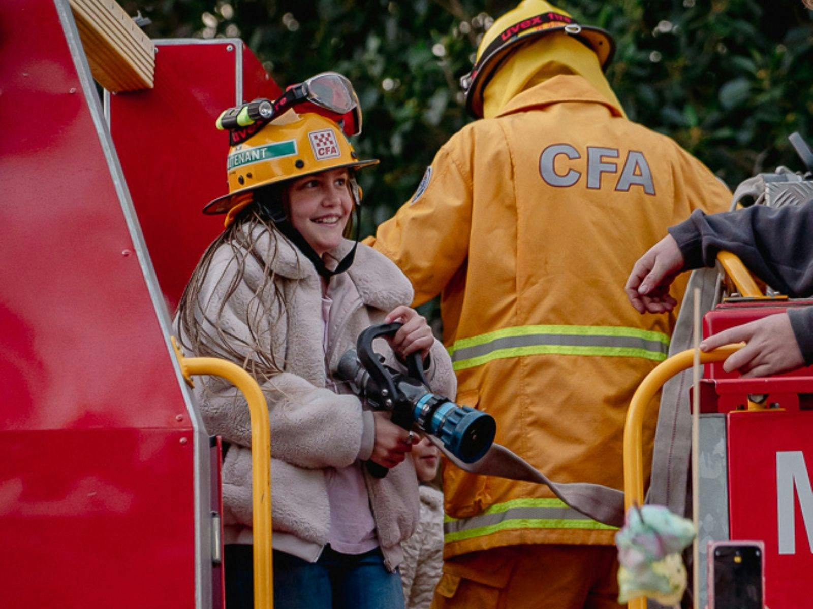 A smiling girl with warm coat and fire hat standing on the back of a fire truck holding fire hose