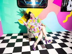 A woman flings her long dark hair while seated. The walls behind her are colourful as is her dress.