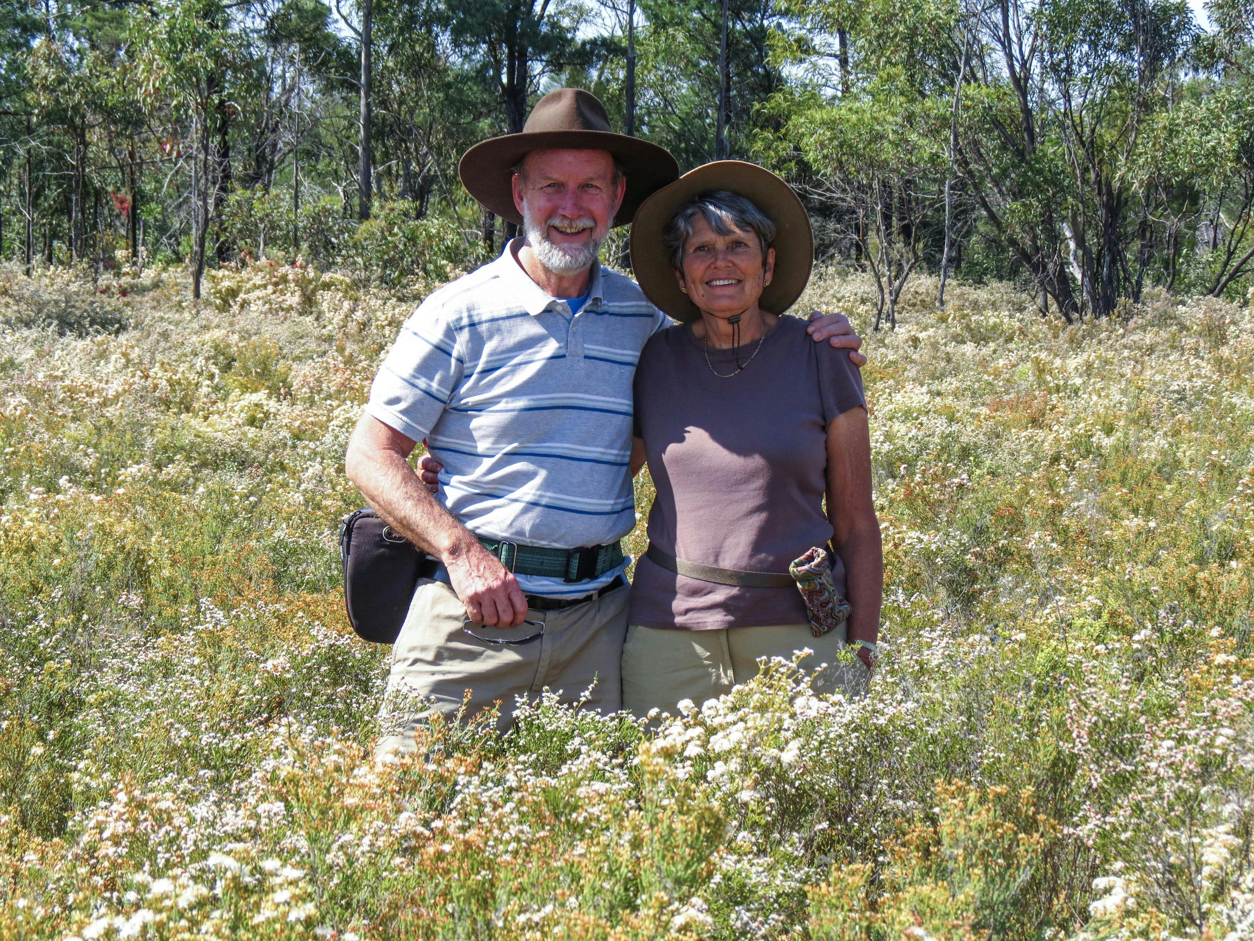 An elderly couple standing in tall grass surrounded by flowers