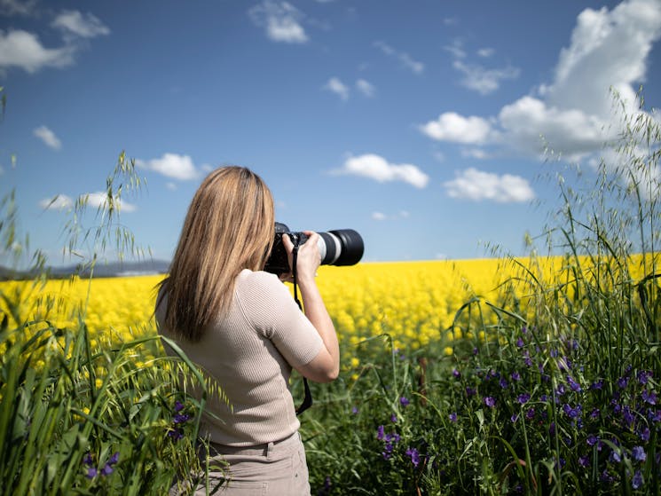 A young photographer stands in front of a beautiful canola field, poised with camera in hand.
