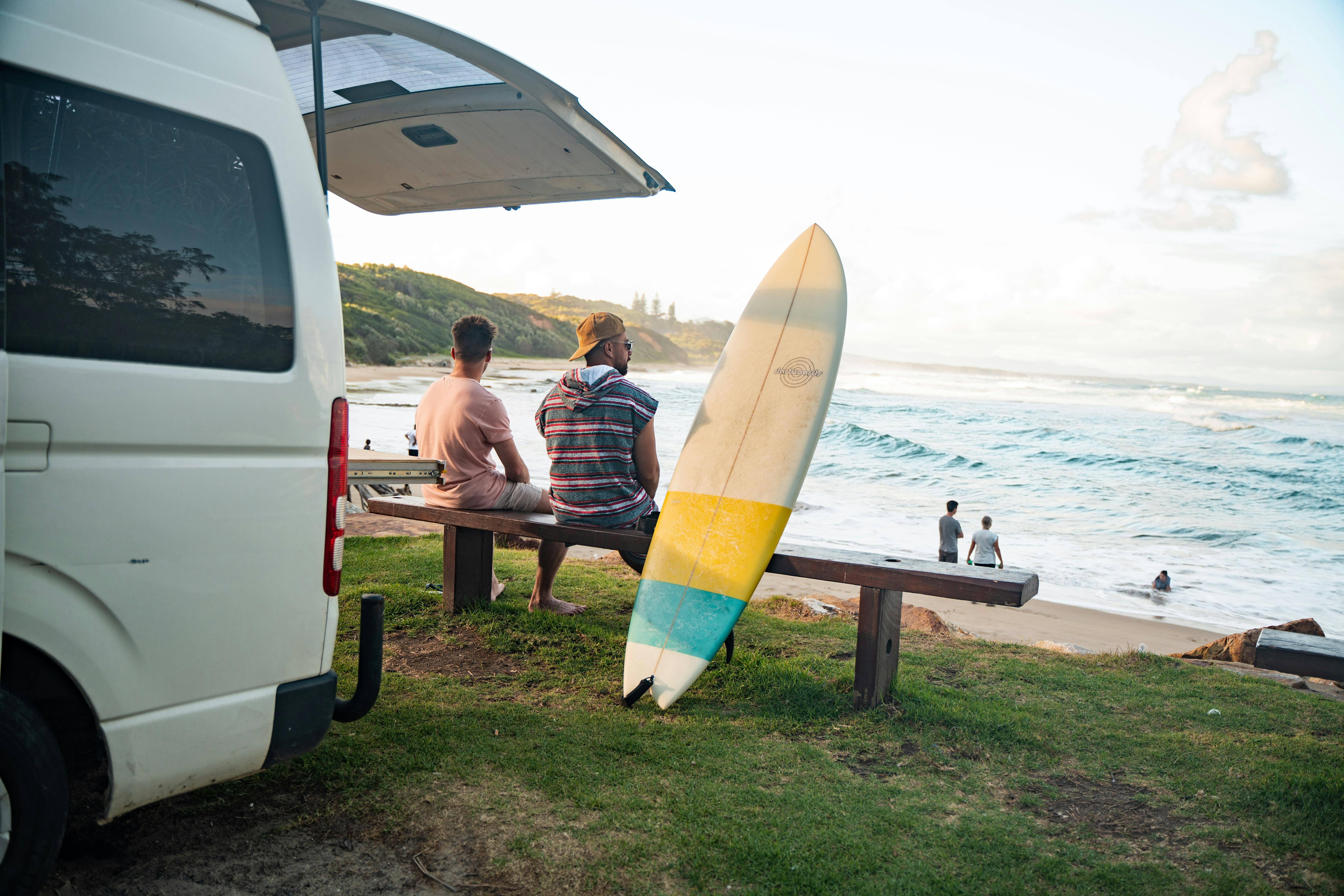 2 men sit on front of their campervan, looking out at the beach