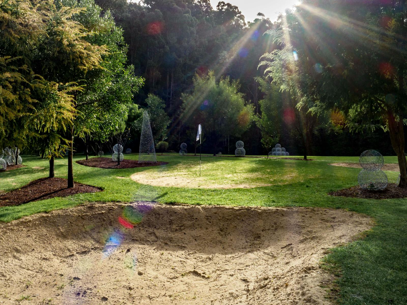 Sand bunker, wire sculptures made onsite, sunlight filtering through tree tops, lush green grass