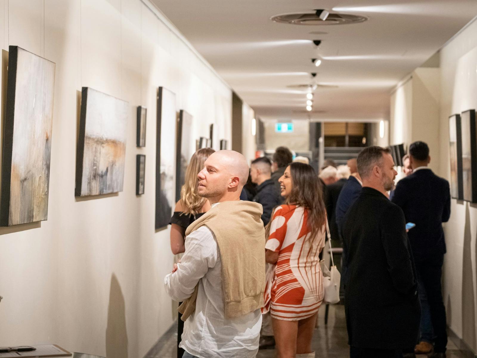 Visitors mingle in a bright gallery corridor, studying framed paintings on white walls