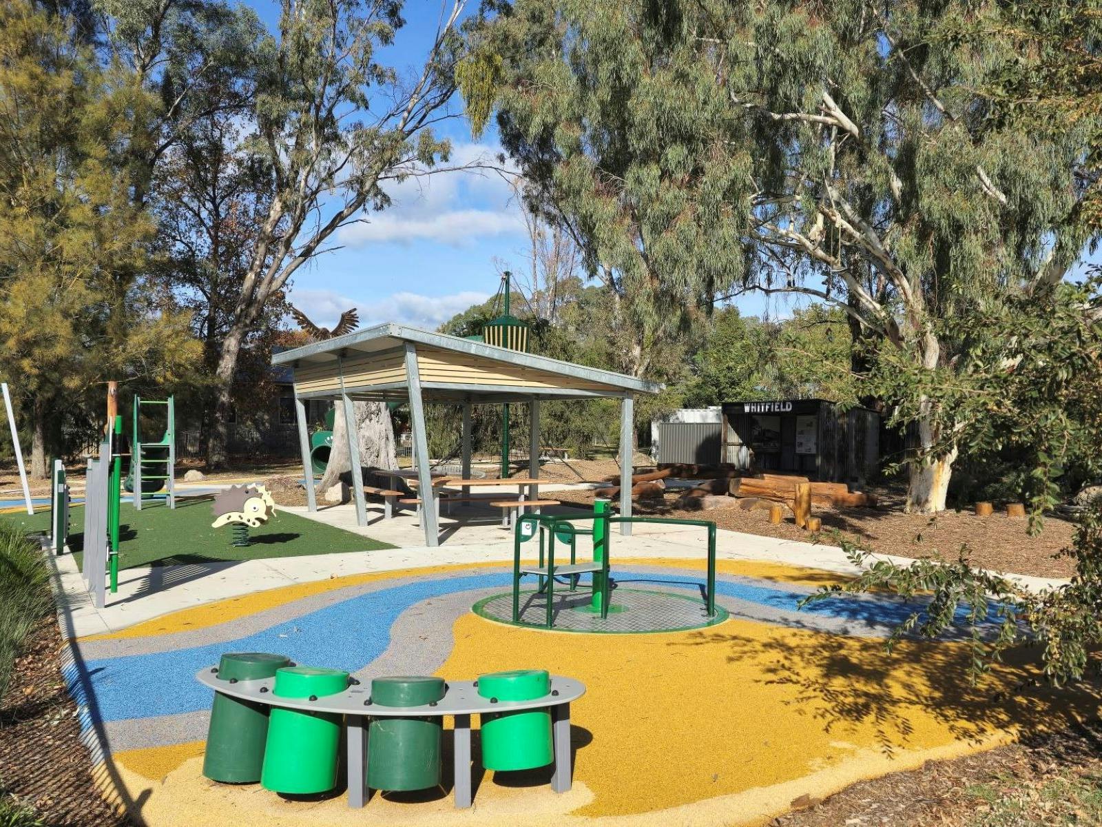 Playground area with green drums on a yellow surface. Shaded seating area in background