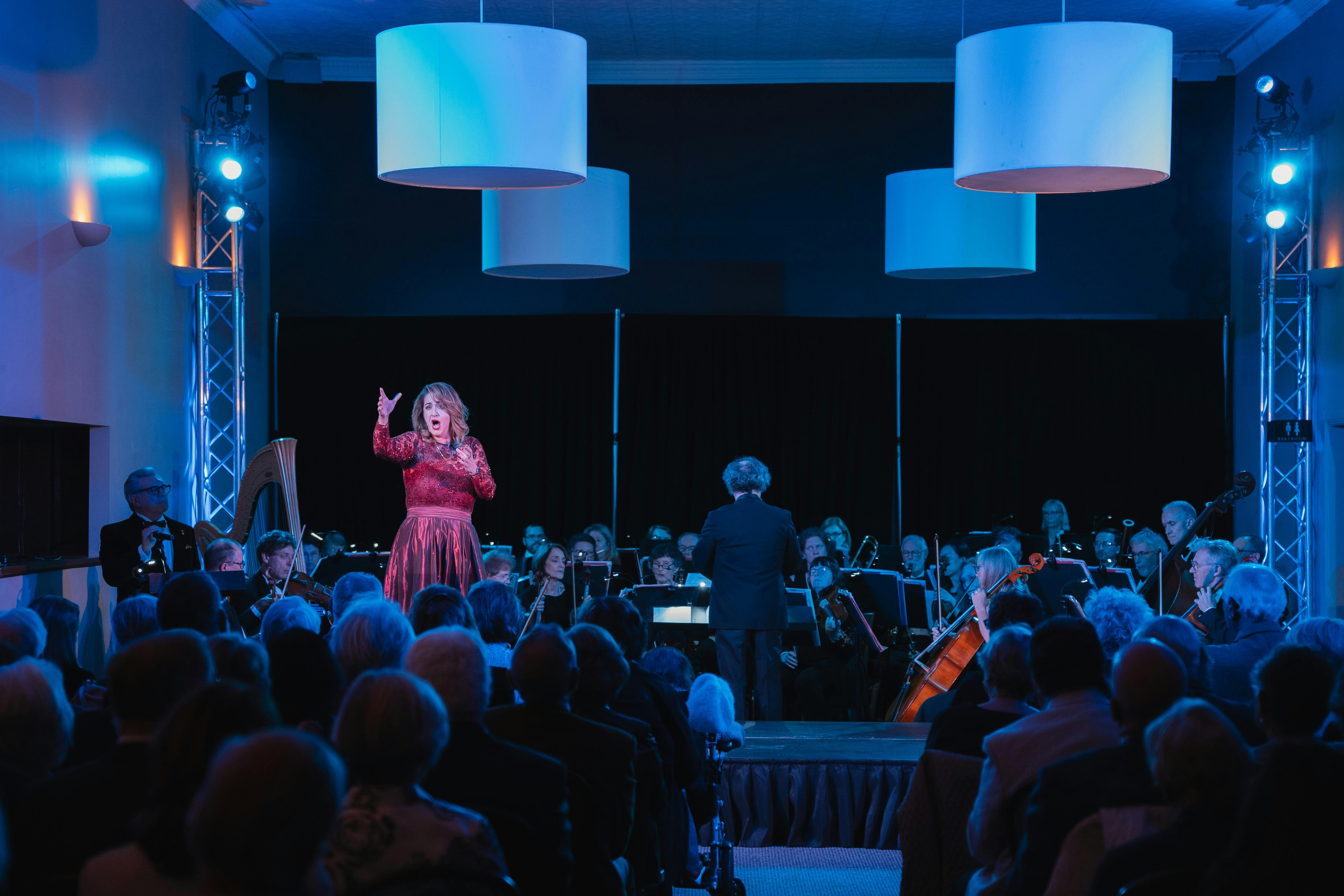 A singer in a red dress on stage singing with her arms up and a full orchestra behind her