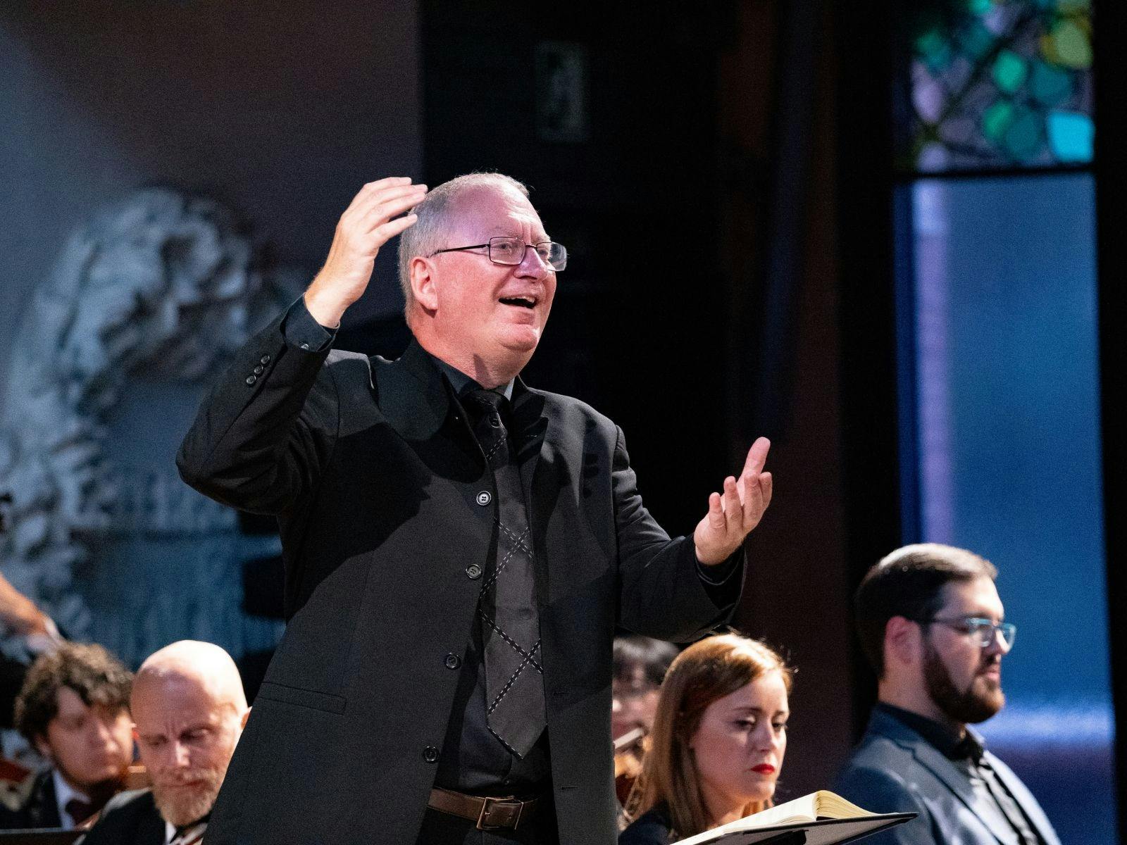 Conductor in formal attire passionately leading a choir and orchestra during a performance.
