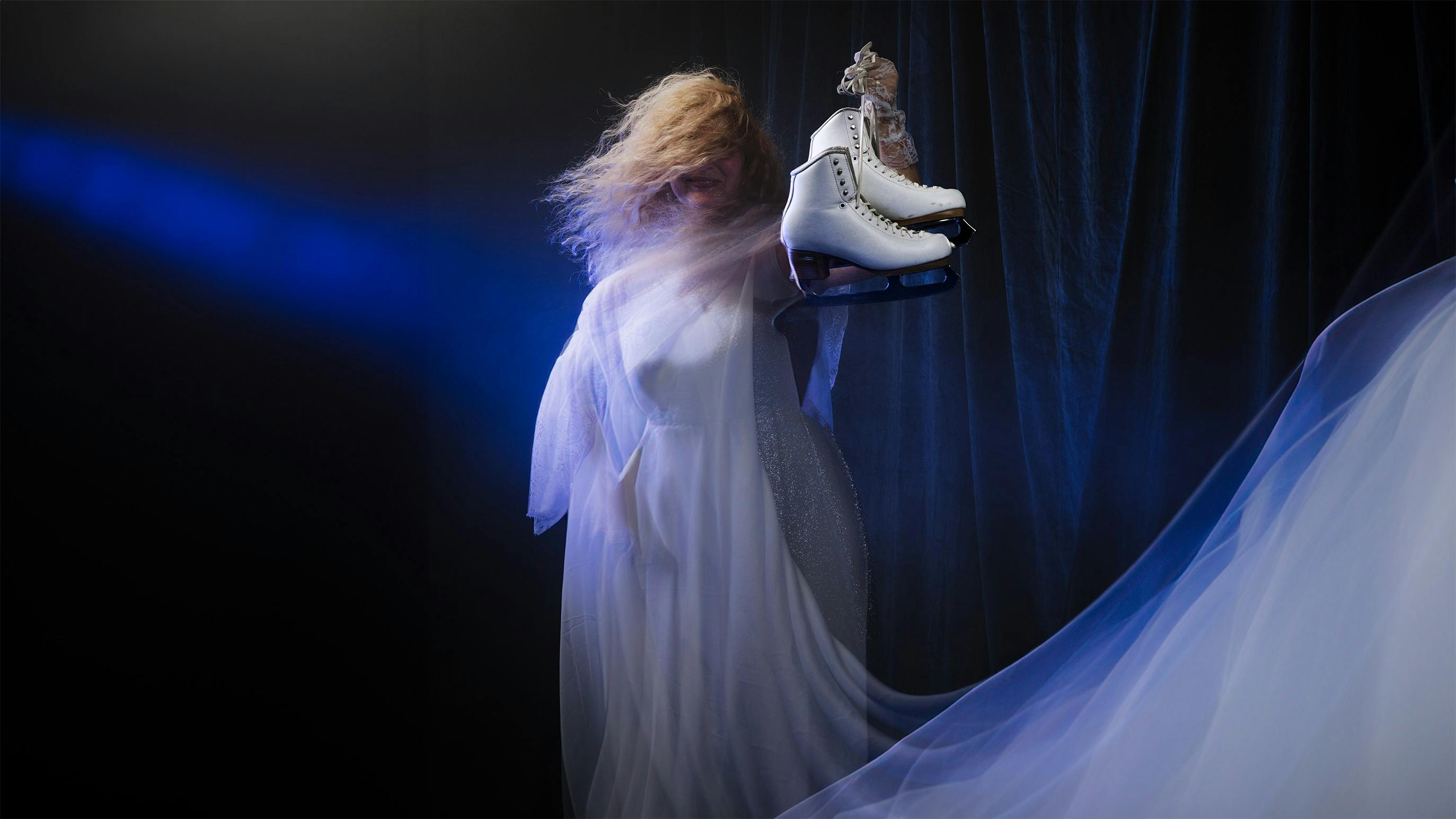 A woman with long blond hair wears a white flowing dress and holds a pair of white skates