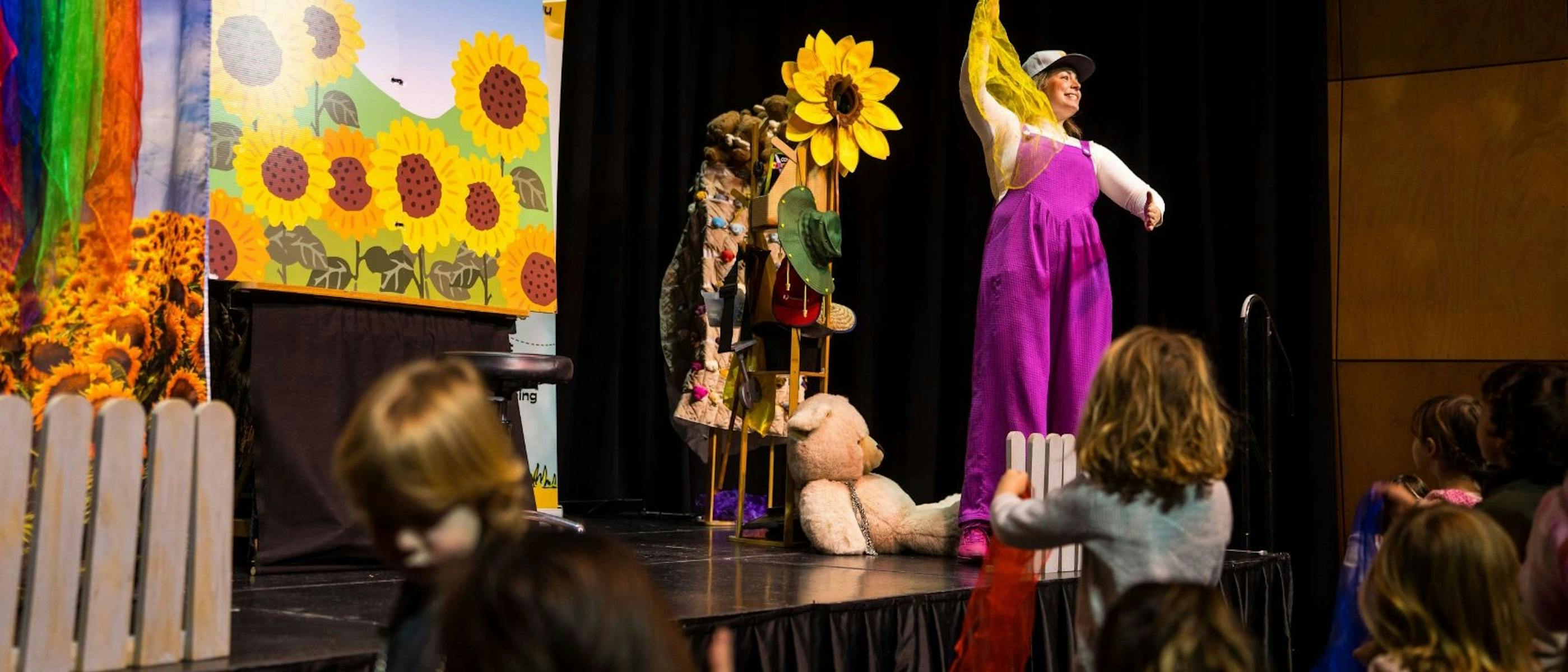 A woman in pink overalls performs on a stage with toys while children watch