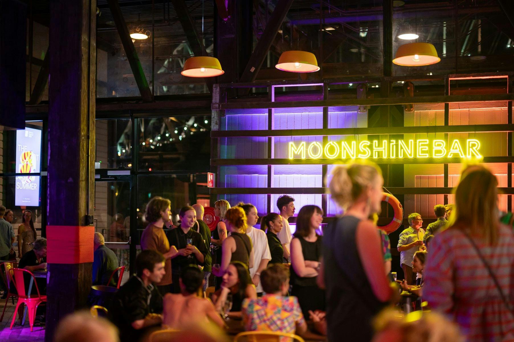 A lively crowd enjoying drinks at a bar adorned with vibrant neon moonshine bar sign