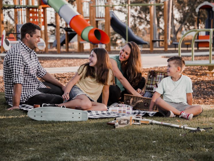 Family enjoying picnic whilst sitting on th elawn with huge playground in the background