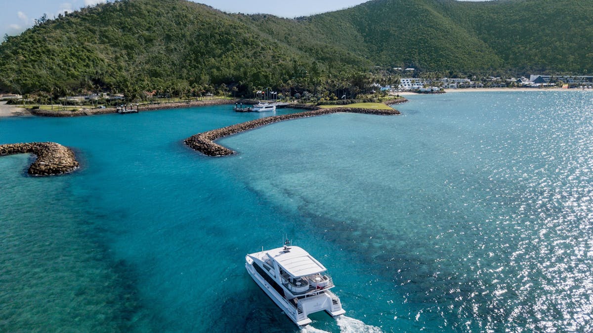 A white boat sails toward Hayman Island, featuring turquoise waters, rocky inlets, and green hills.