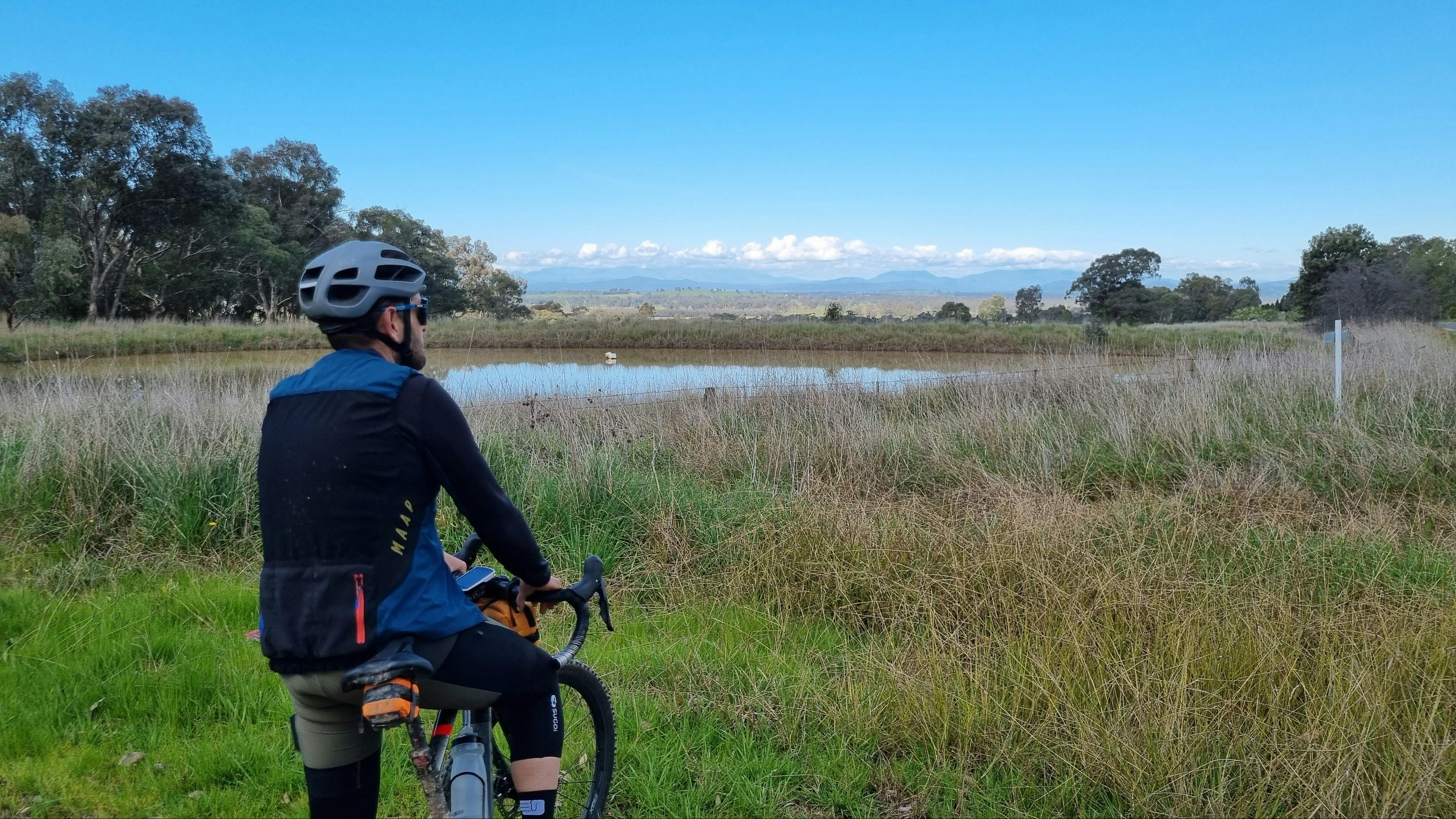 Cyclist stopped looking over the view, grass, dam distant trees, background of mountains, blue sky