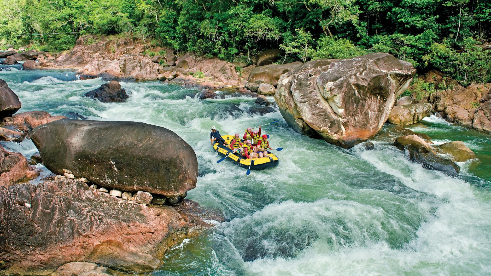 Wide shot of a rafting boat surrounded by blue water, lush rainforest and large boulders in Tully Wide shot of a rafting boat surrounded by blue water, lush rainforest and large boulders in Tully