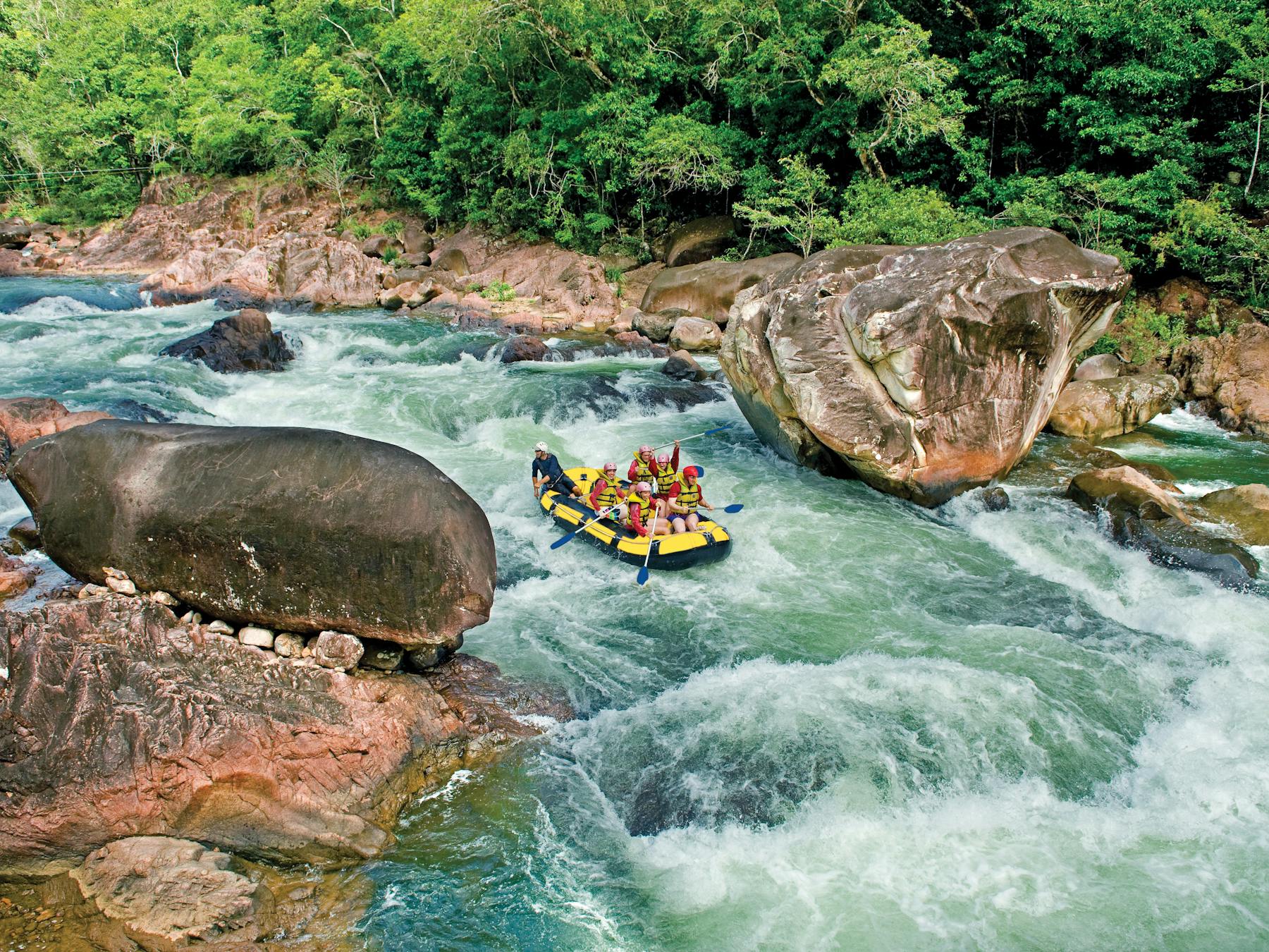 Wide shot of a rafting boat surrounded by blue water, lush rainforest and large boulders in Tully