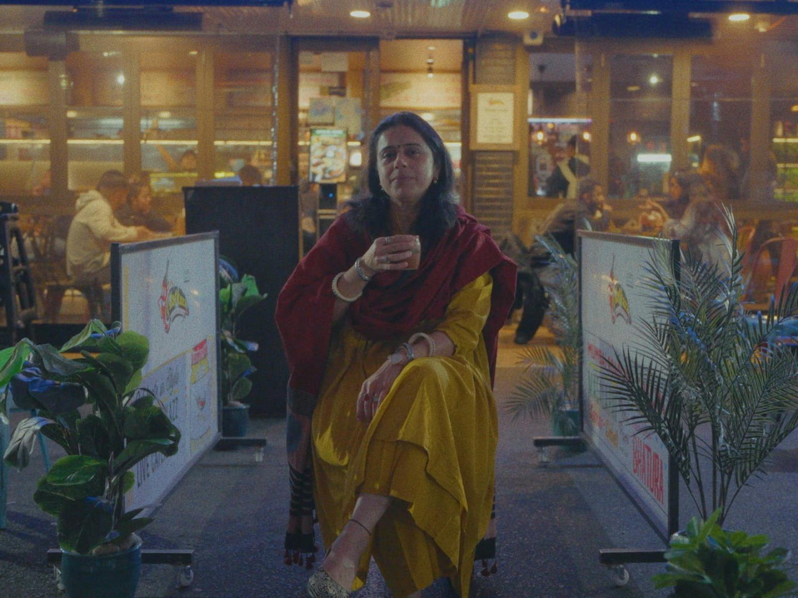 A South Asian woman in a mustard yellow sari and a burgundy shawl with a cup of tea in her hand