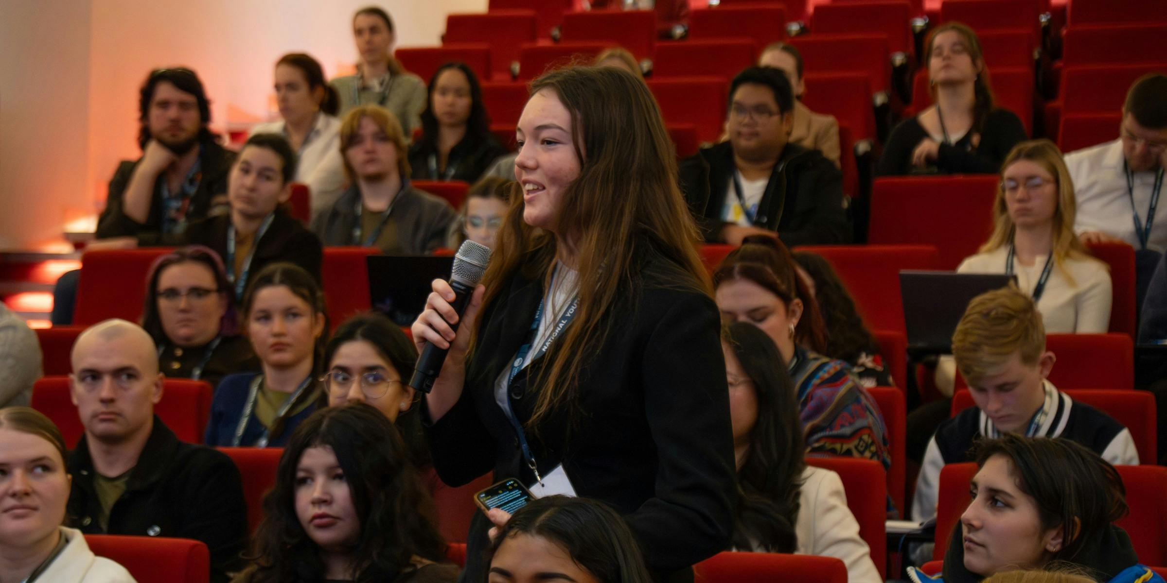Photo in theatre, in focus is a conference attendee with a microphone asking a question
