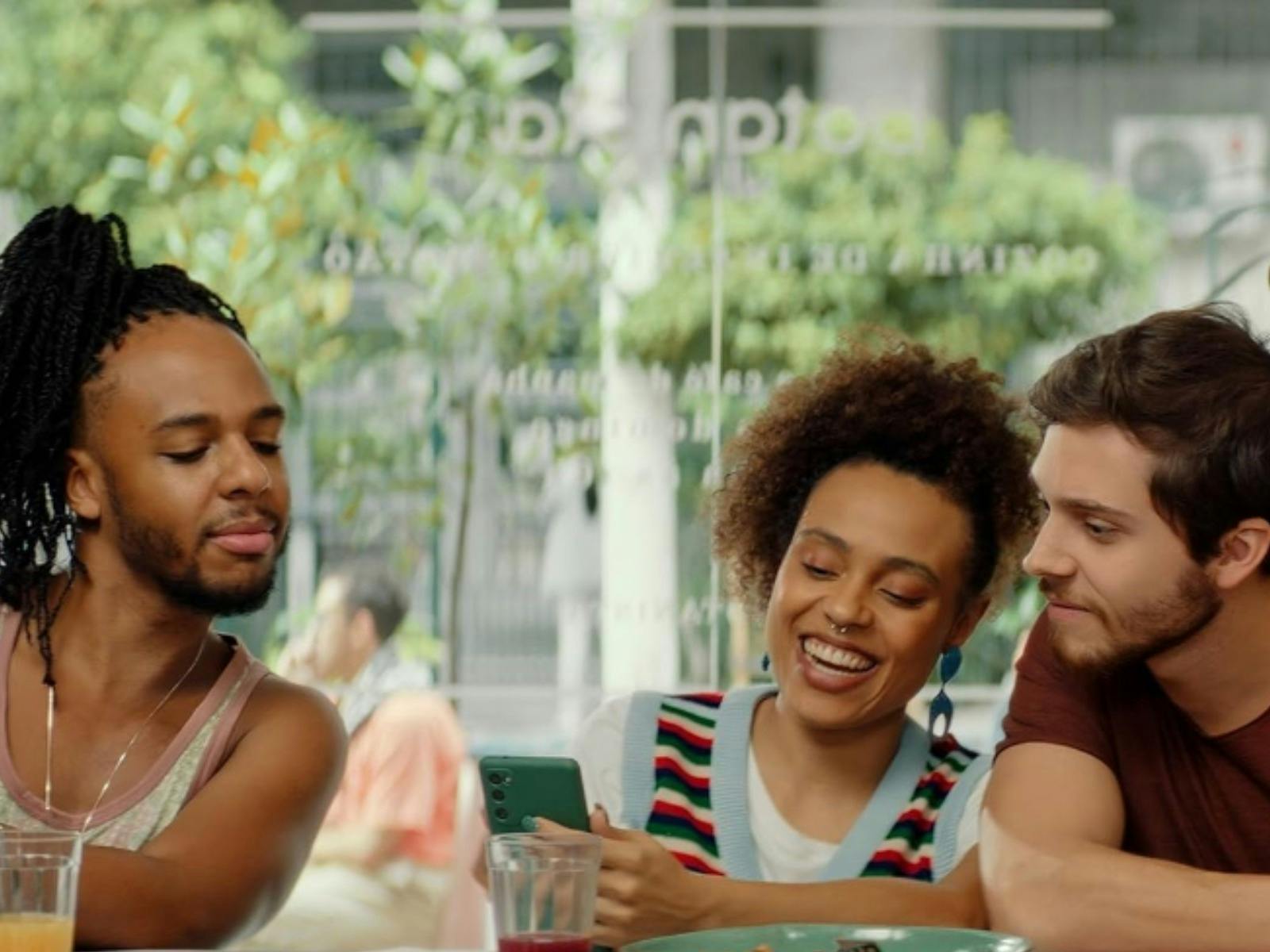 Three friends in a cafe. They each have a phone in their hand and are looking at one friend's phone.
