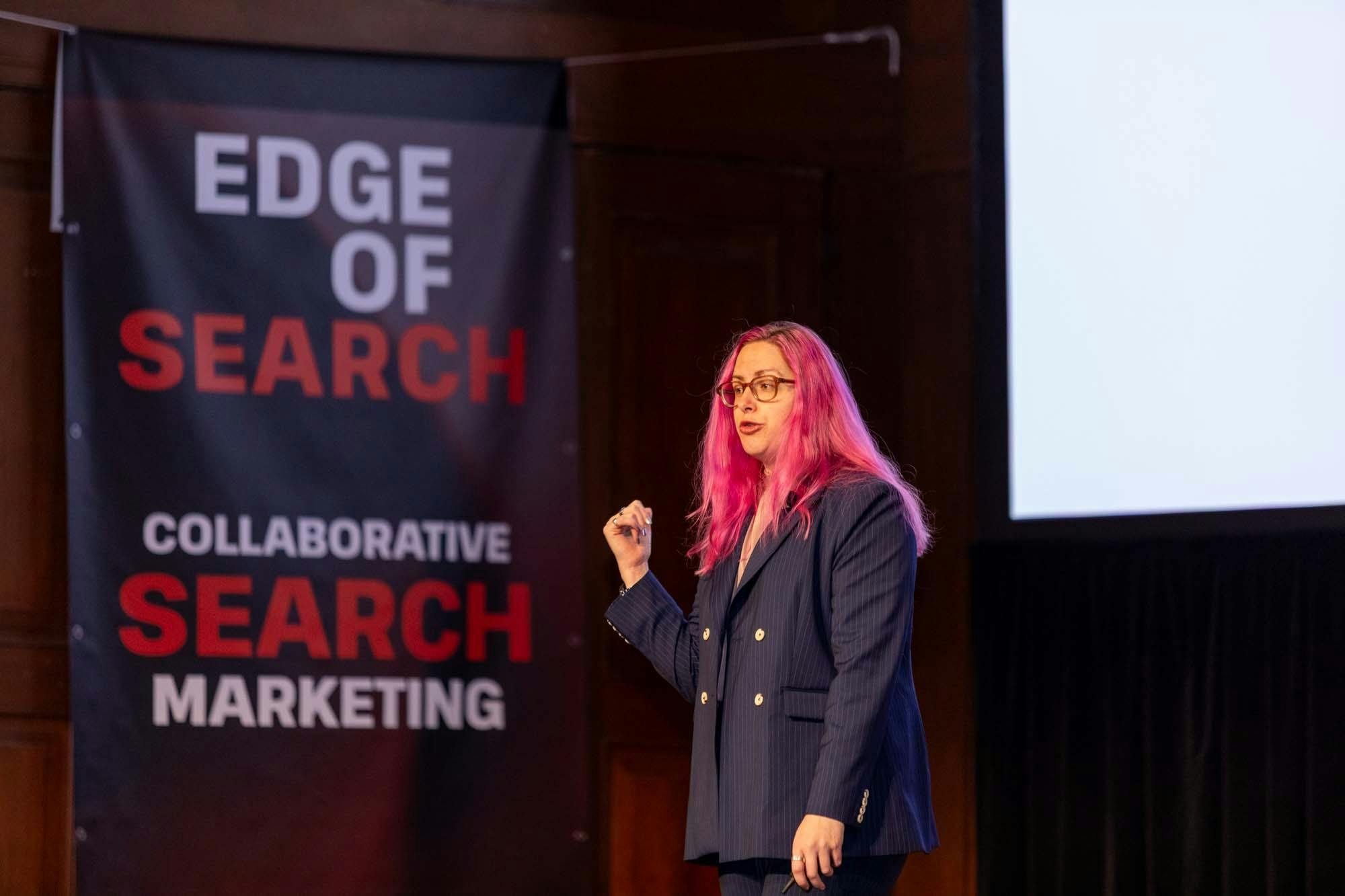 Woman with pink hair in a blue suit talking on stage in front of black, white and red banner.
