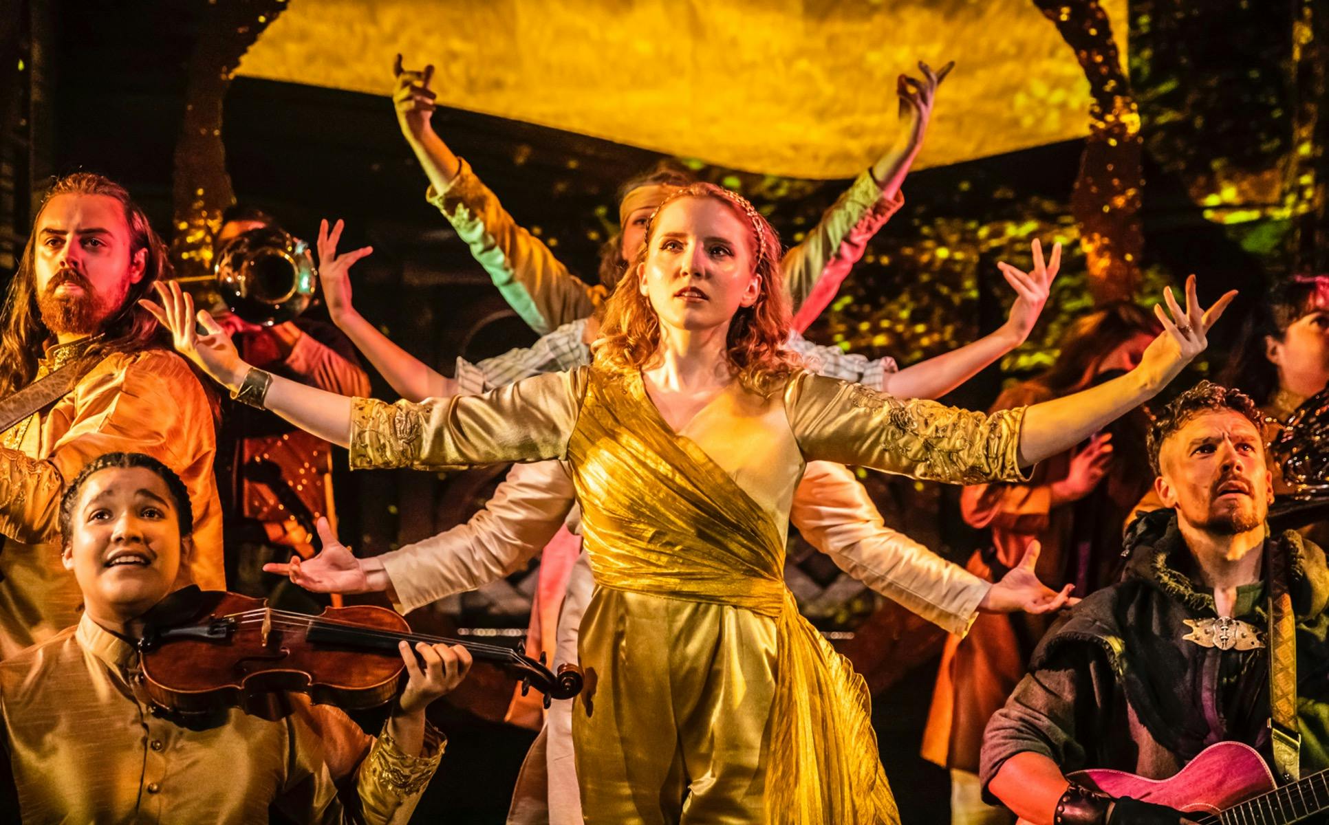 A woman with red hair and a gold dress stands with her arms outstretched. She is surrounded by music