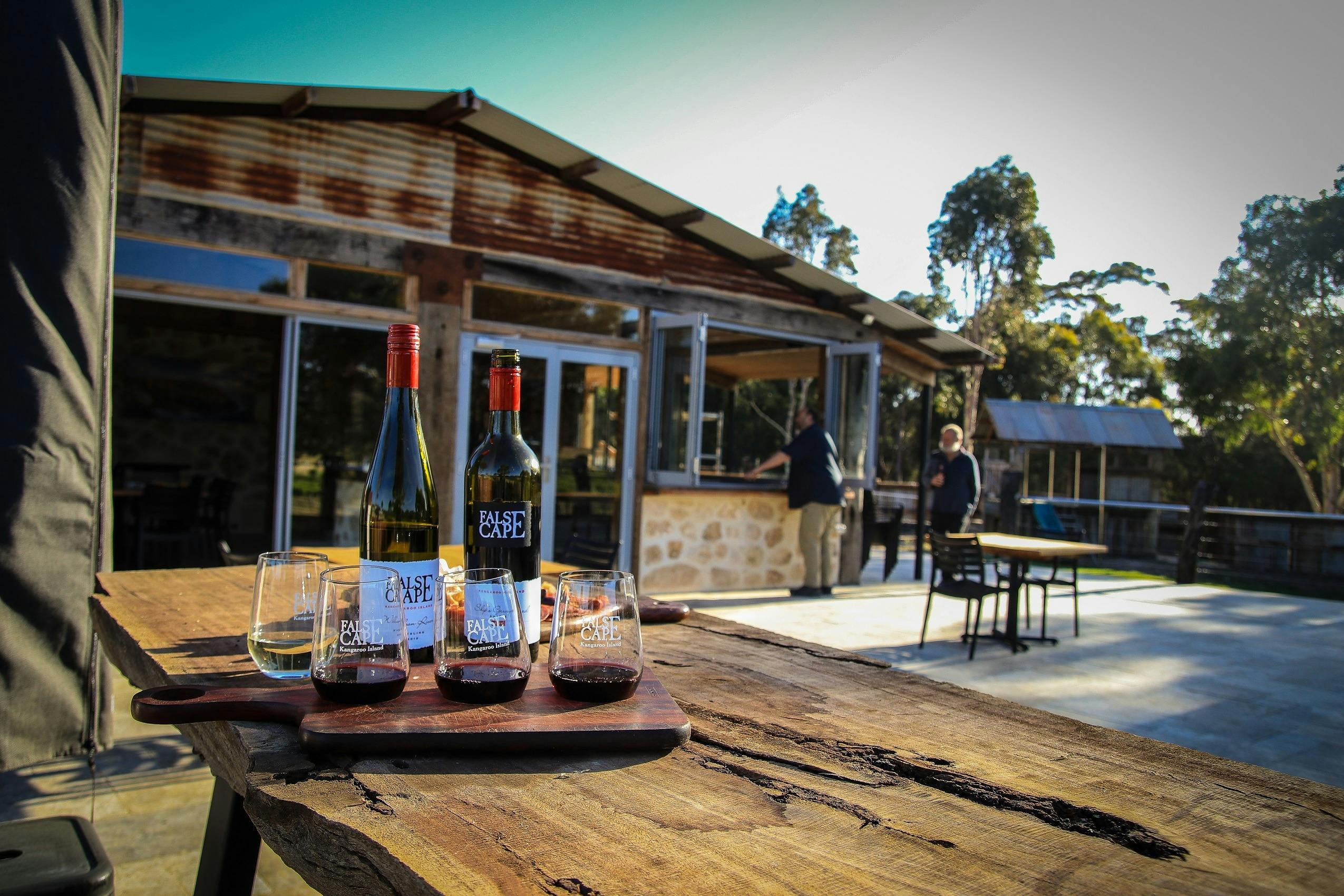wine tasting flight in foreground with two bottles of wine and cheese board, and 2 men in background