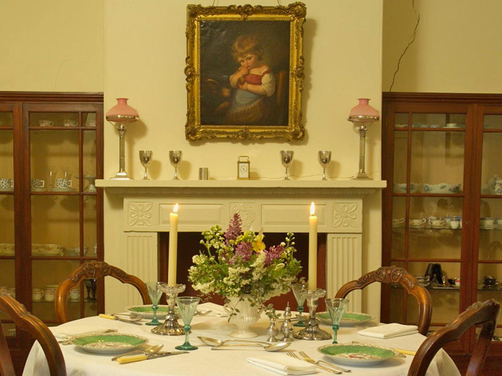 A dining table is set with a vase of flowers and lit candles in the middle. A mantlepiece is behind