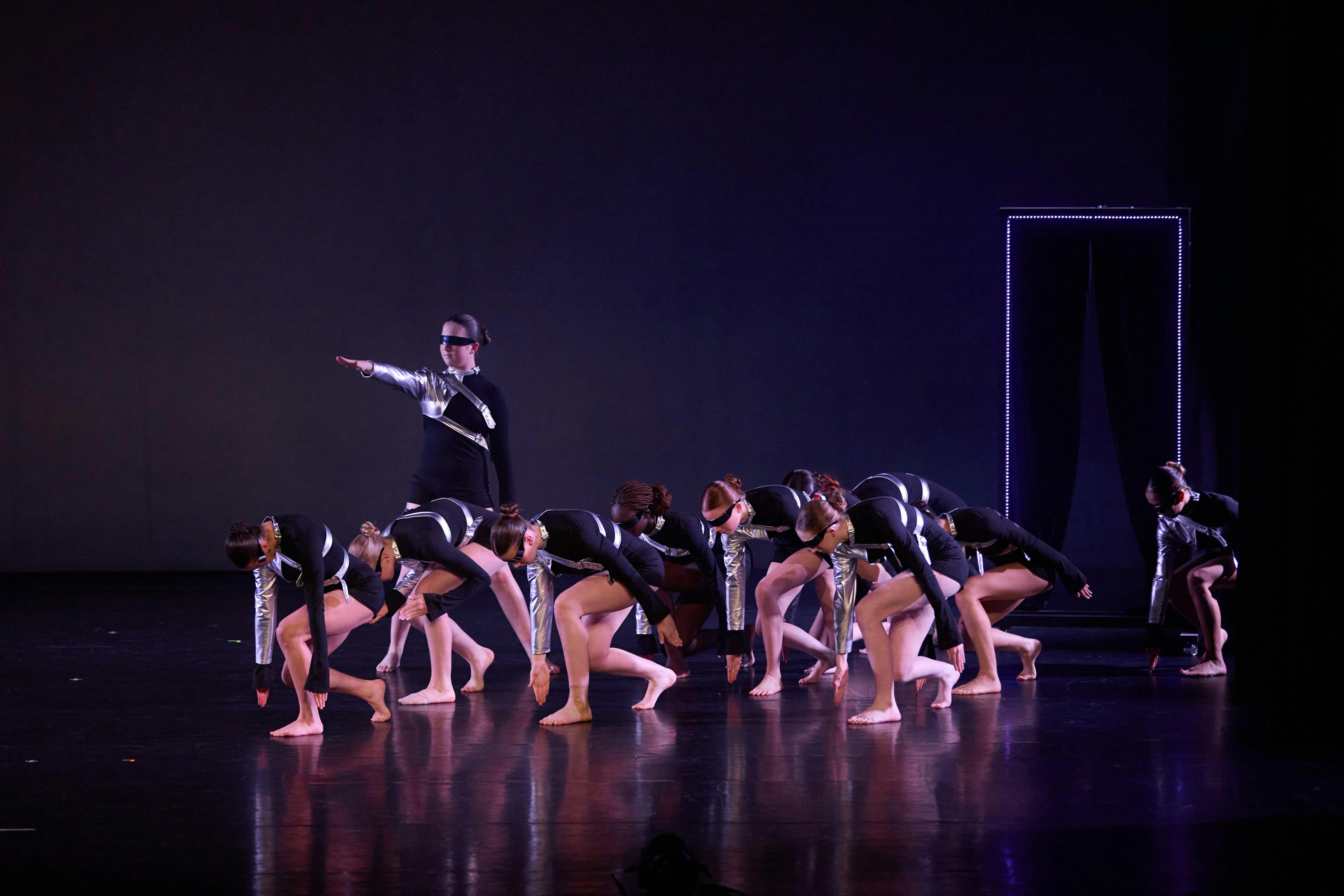 A group of dancers in black costumes performing a synchronized pose under dramatic stage lighting.