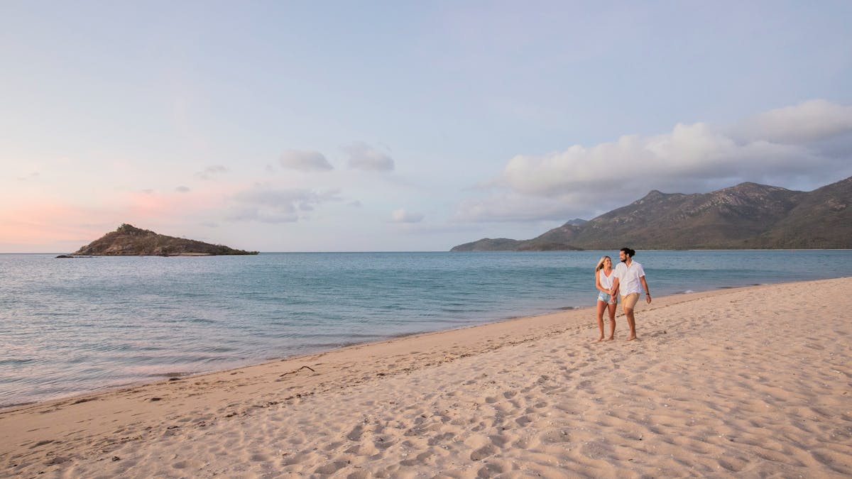 A happy couple walking along the sandy beach during sunset, hilly islands in the background