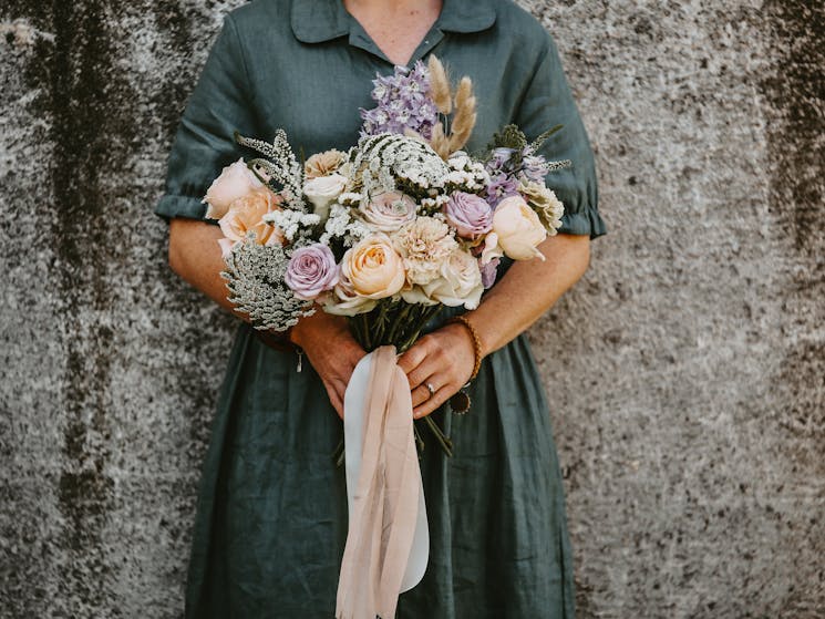 Neck down photo of a lady in a green dress holding a bouquet of pink and purple flowers with ribbons