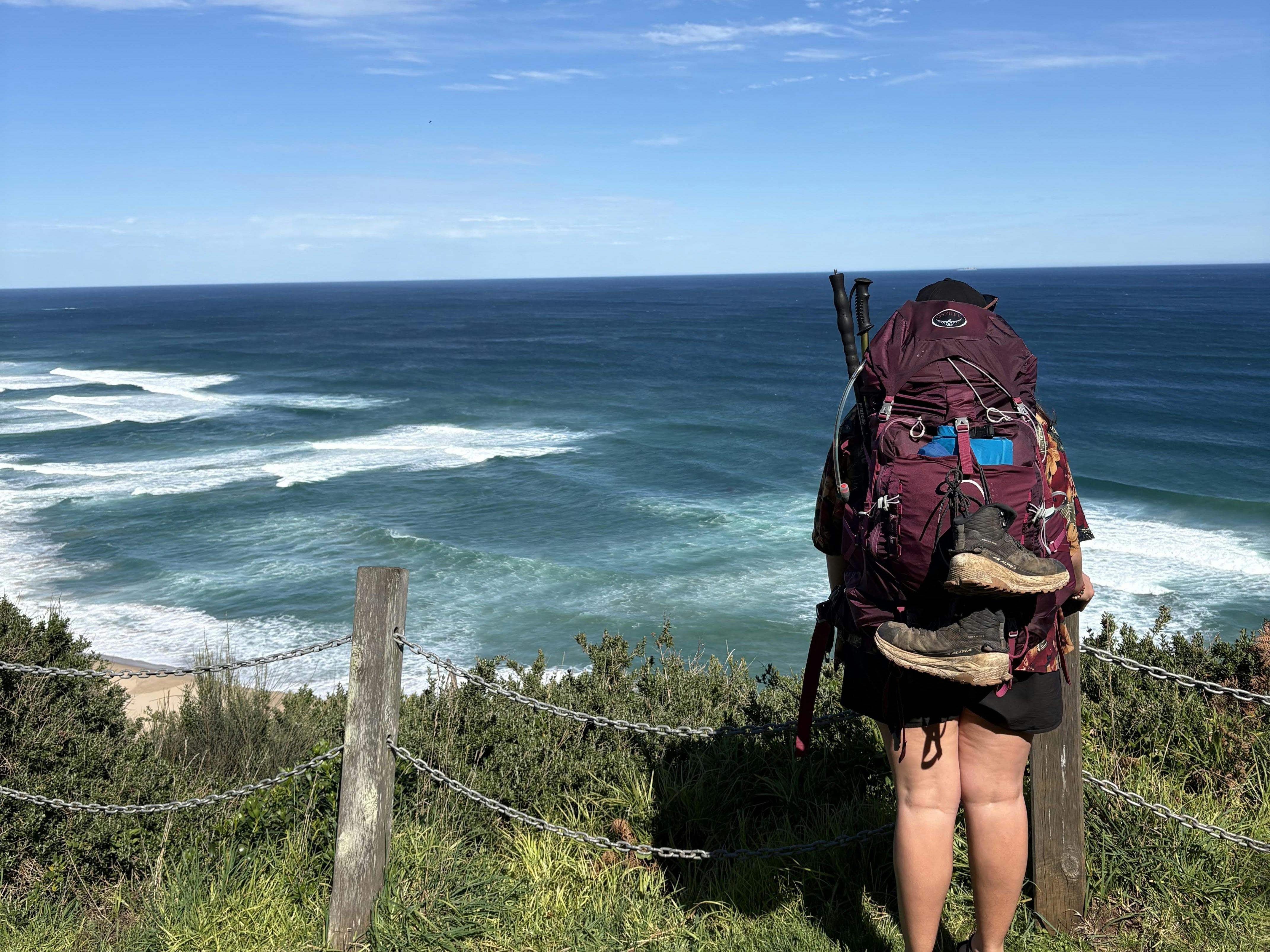 women with hiking pack, looking out to the ocean from a green grasy cliff