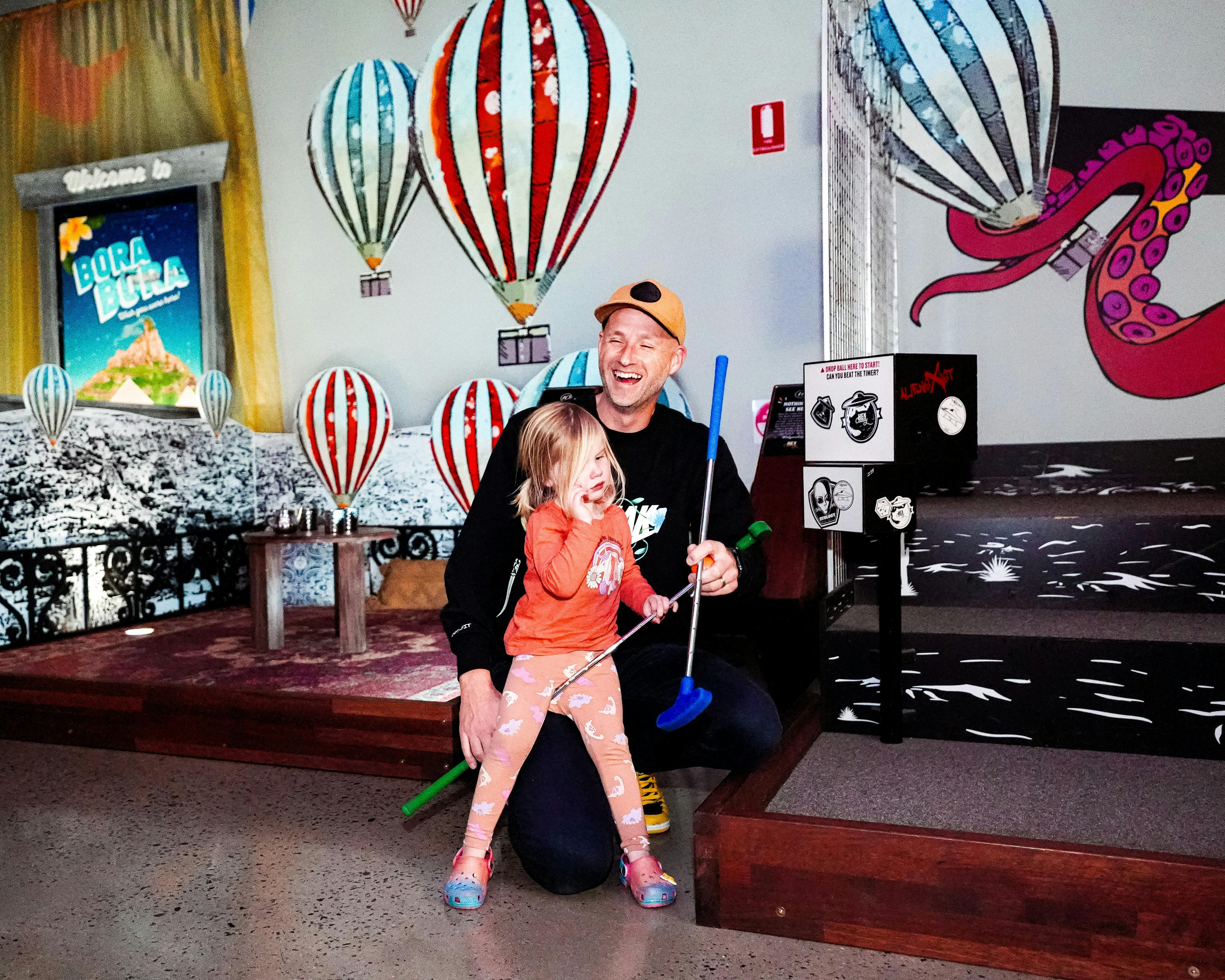 A father and daughter share a laugh while playing indoor mini-golf, surrounded by a colourful theme.