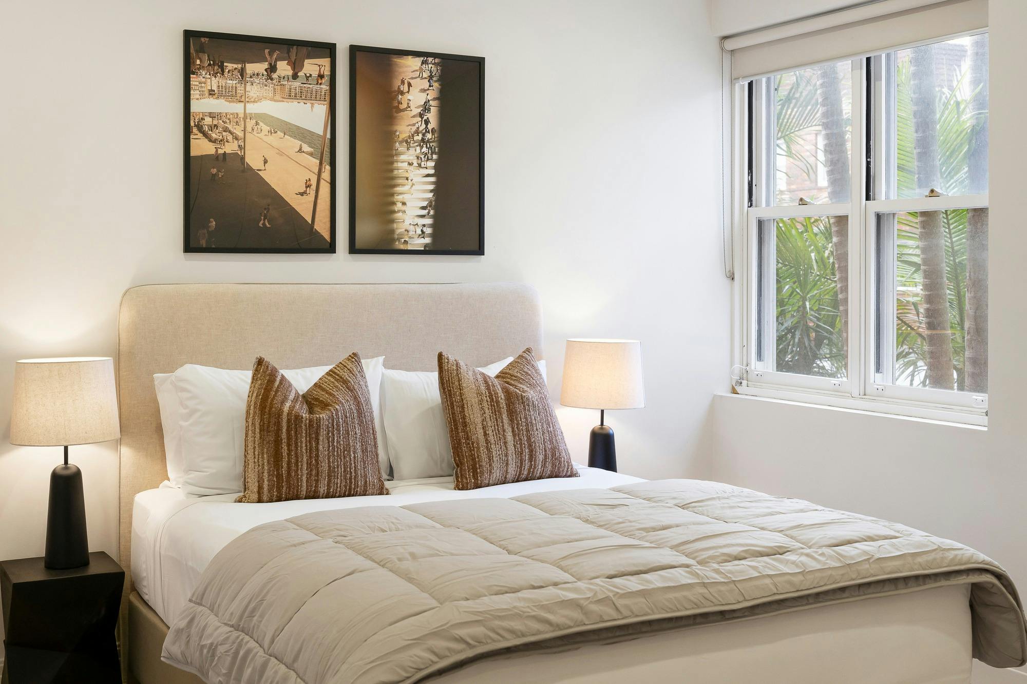 Comfortble bedroom featuring queen sized bed with comforter, large window, framed wall art and lamp.