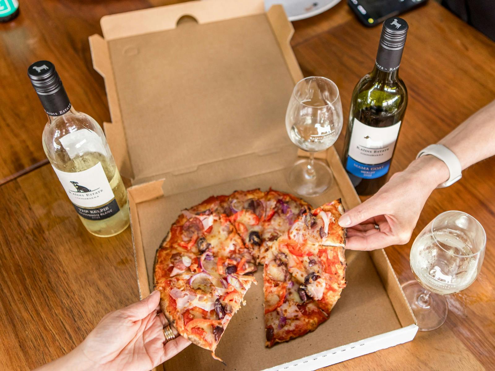 Hands grabbing Raidis pizza from a box with a bottles of Raidis wine on the table.