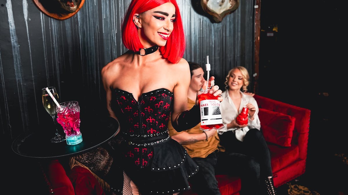 A female waitress in gothic costume serves cocktails to a male and female customer.
