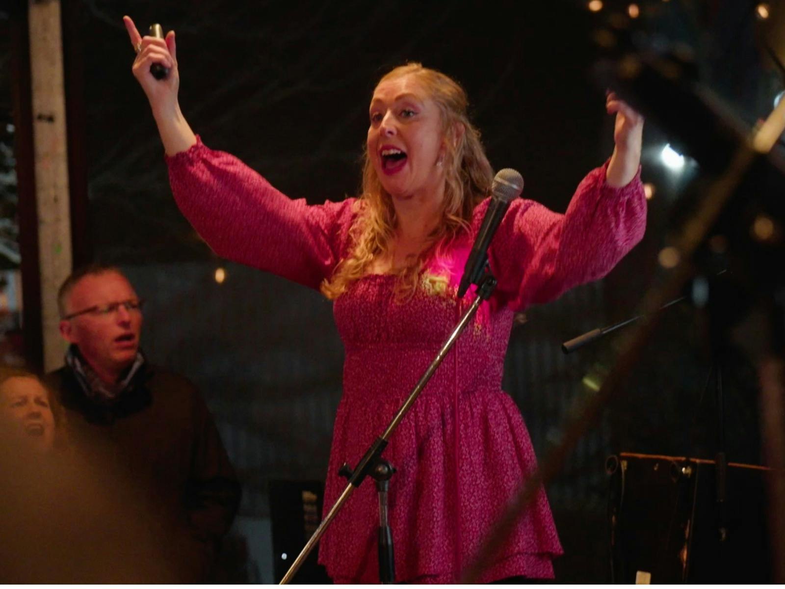 A joyful woman in a bright pink dress sings into a microphone, arms raised, performing energetically