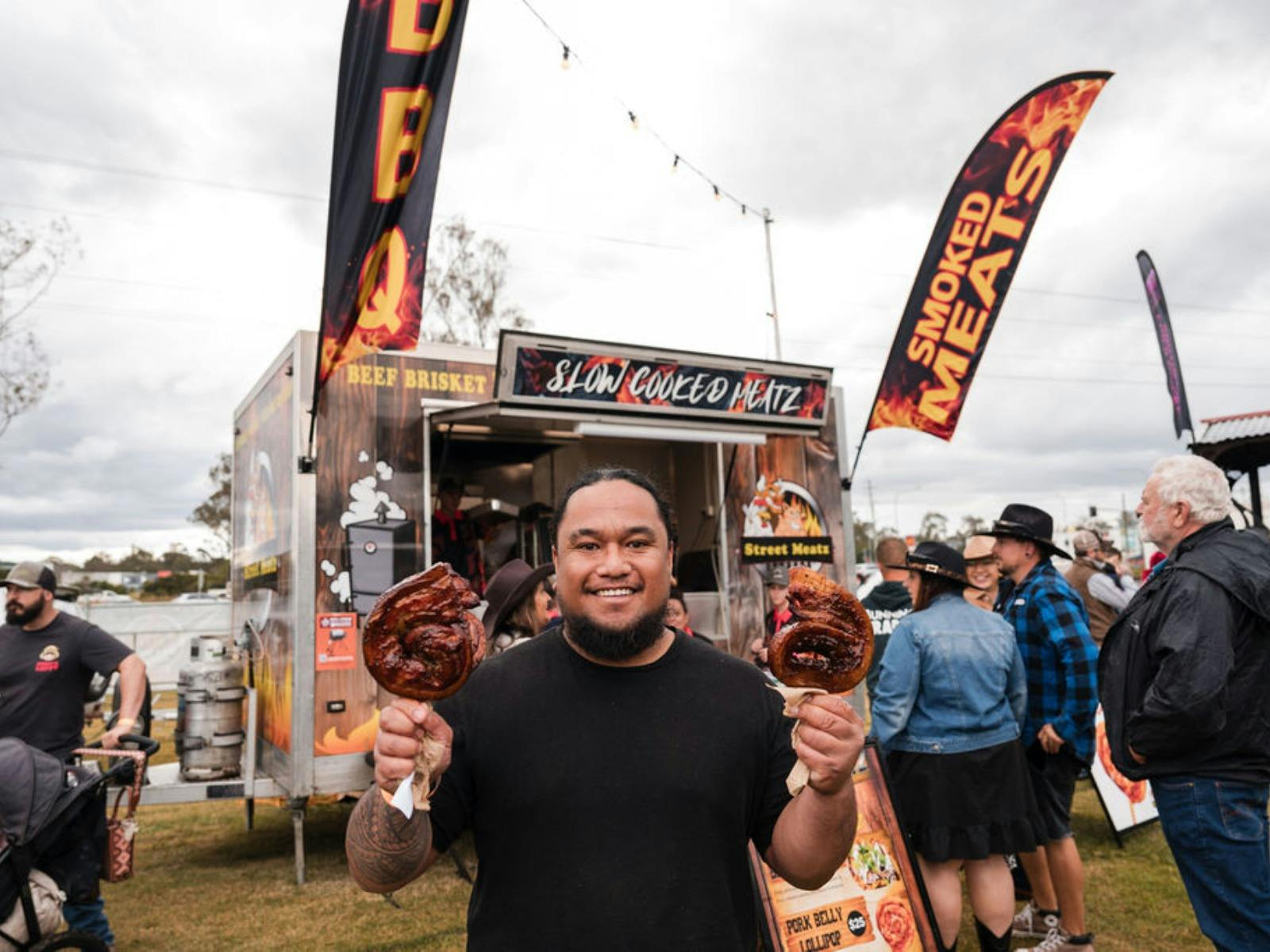 Person holding two smoked meat portions in front of a busy food truck at an outdoor festival.