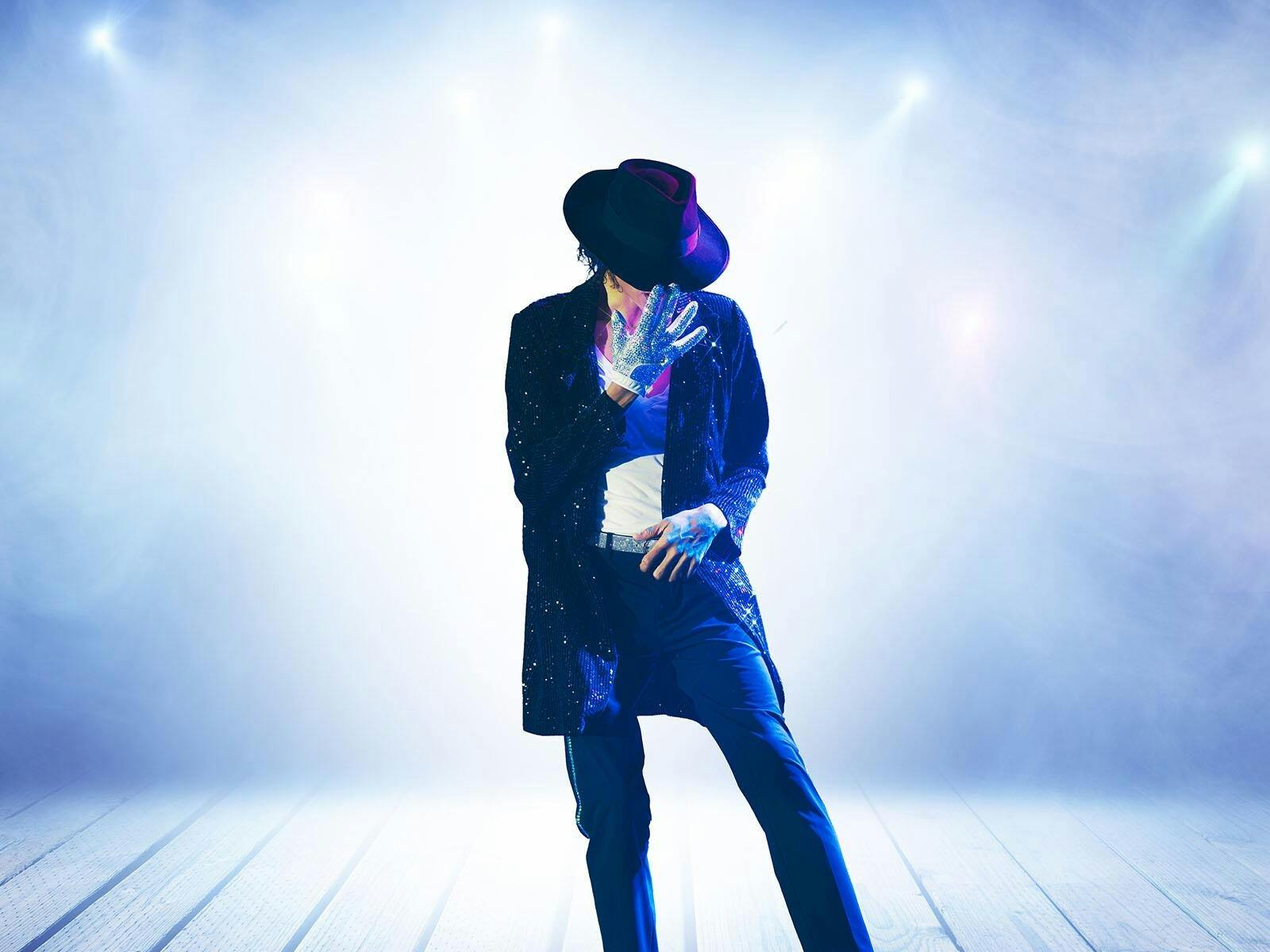 A man in a black jacket and pants with white gloves and a dipped hat stands in front of a white back