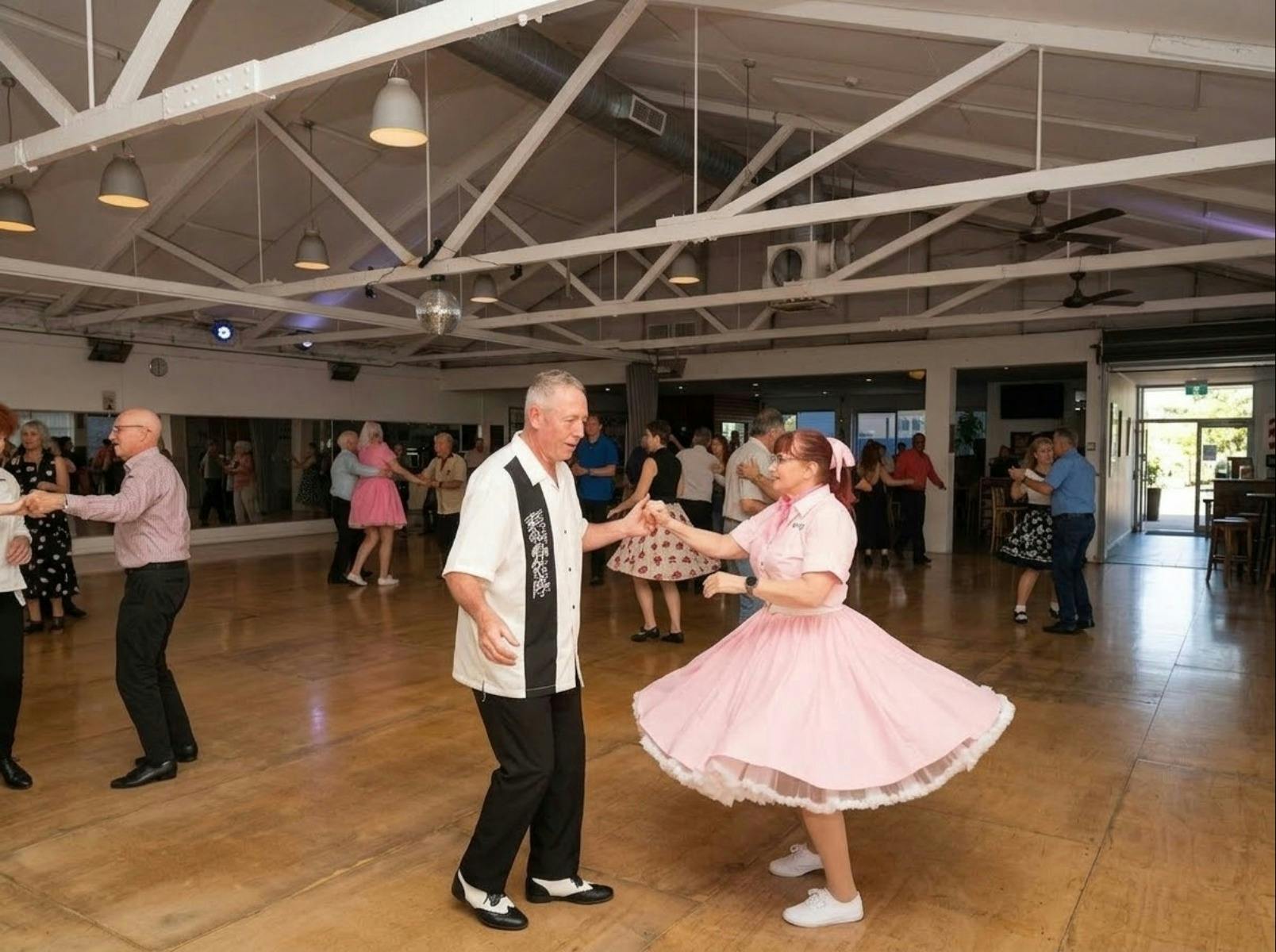 A couple wearing rock'n'roll outfits dances together, other couples in the background enjoy dancing.