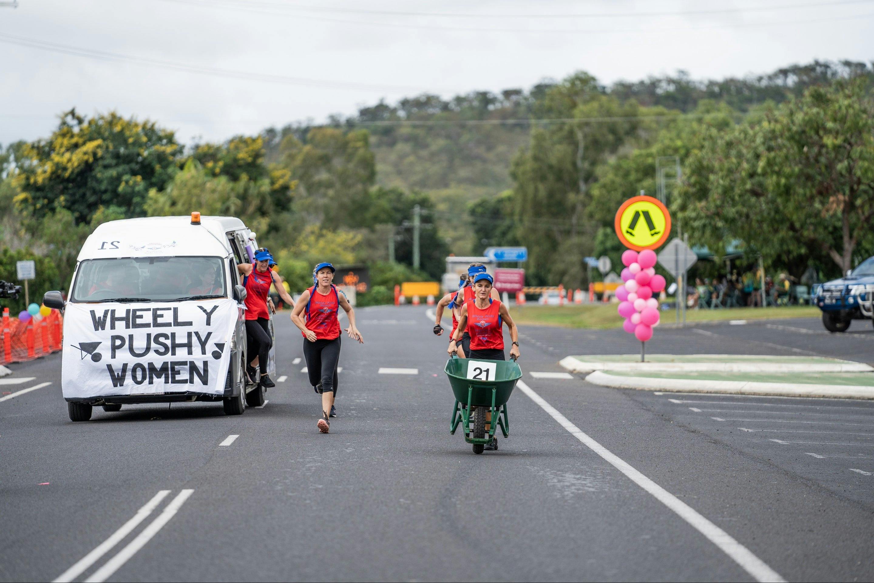 Women in red shirts run, pushing a wheelbarrow, as a van labeled 'Wheely Pushy Women' drives beside.