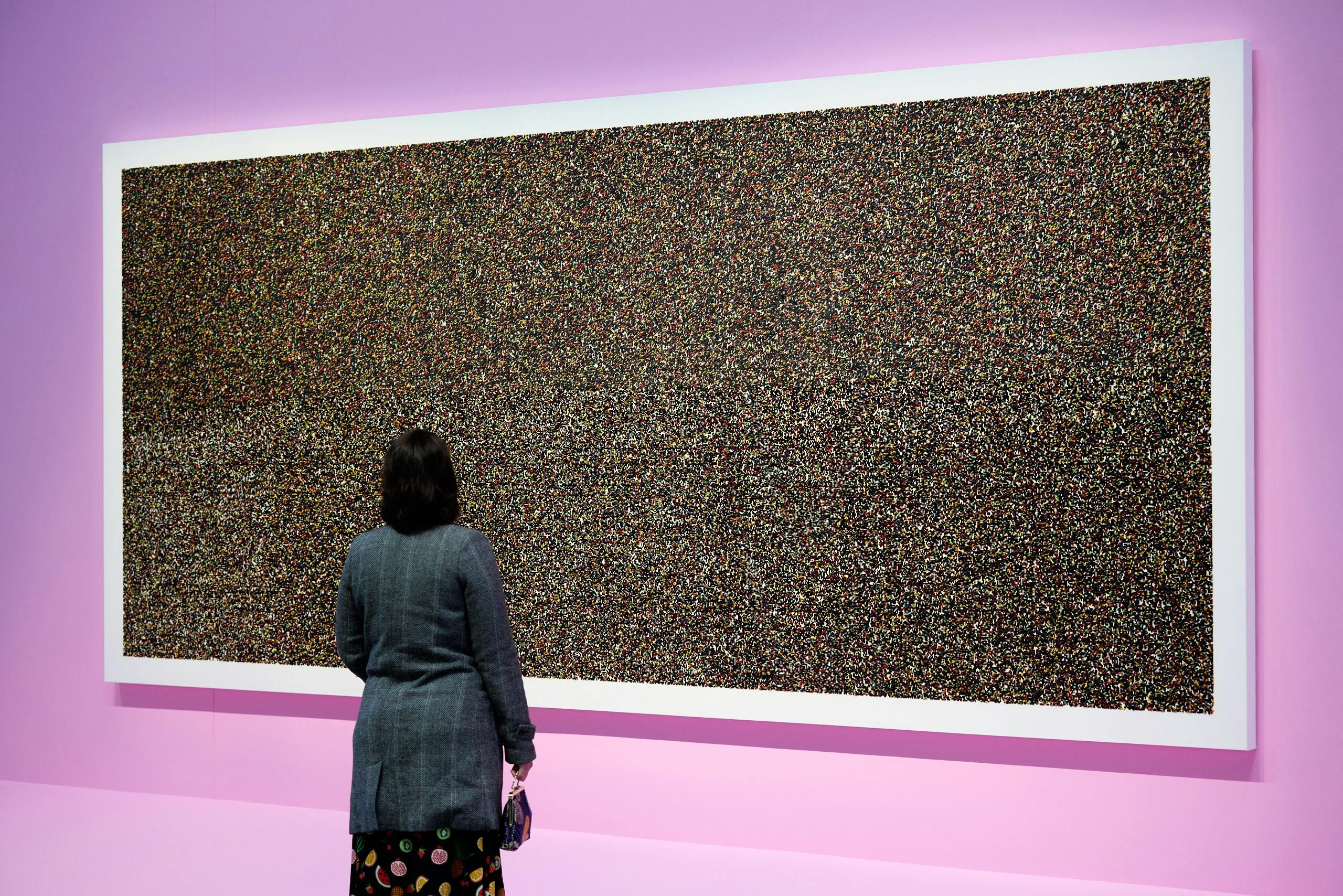 A person stands in front of a bright pink wall on which hangs a large multi coloured painting