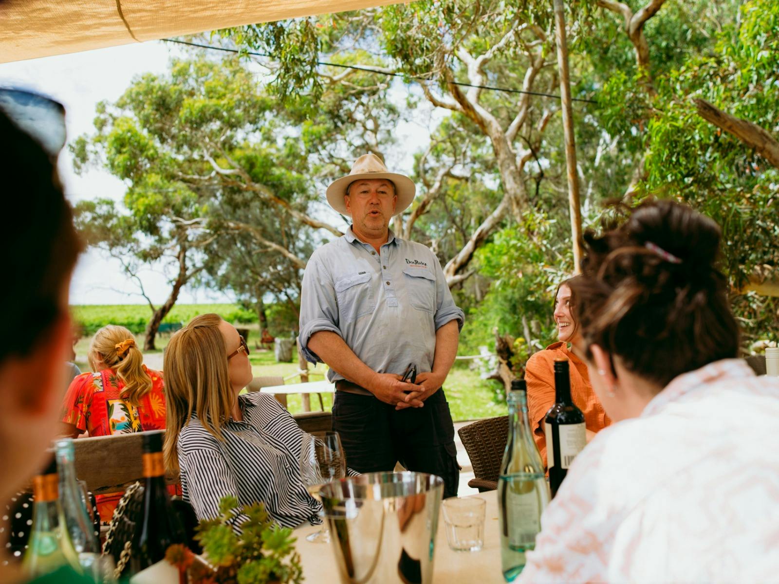A group of people seated outside around a table looking up a man standing beside them